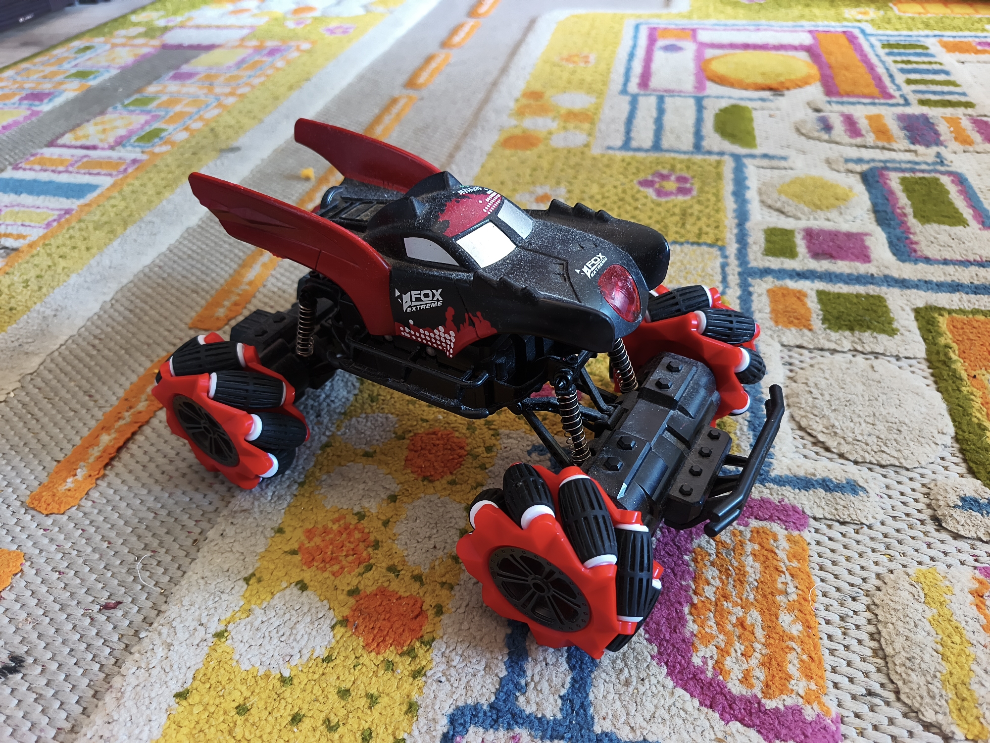 A black and red remote-controlled stunt car with oversized, multi-directional wheels is positioned on a colorful, patterned rug. The car has a futuristic design with prominent red wing-like spoilers and the "FOX EXTREME" logo visible on its body. The rug features geometric shapes and bright colors, including yellow, orange, pink, and blue. The scene appears to be an indoor setting, possibly a child's playroom or living room. There is no information available to indicate the location within the city of Baranavichy, Belarus.