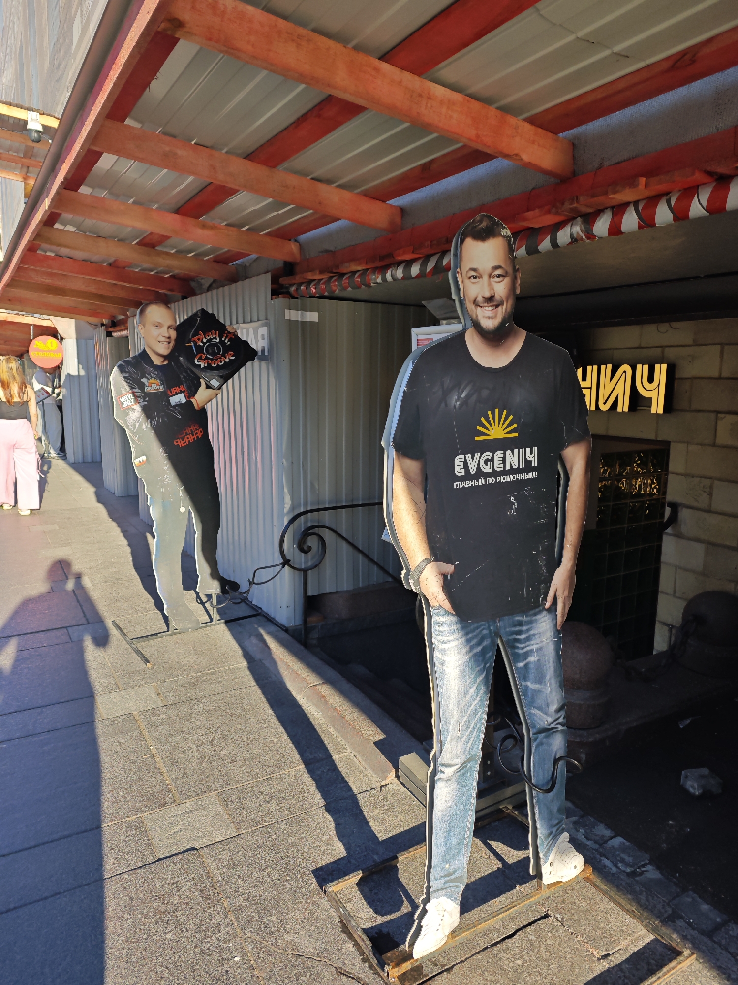 The image depicts an outdoor setting, likely a street or public space, with a cobblestone pavement. Two life-sized cardboard cutouts of men stand on the pavement, each supported by metal stands. The cutout on the left is of a man in a black leather-like jacket and trousers, holding a black record with red and white text that reads "Play it Groove." The cutout on the right is of a man in a black t-shirt with yellow graphics and text that includes "EVGENIY" and "главный по рюмочным," and blue distressed jeans. Both cutouts are positioned under a wooden and metal canopy structure. In the background, a building with brickwork and illuminated signage with Cyrillic letters is visible. A woman wearing pink trousers and a black top is walking away in the distance on the left. The scene is bathed in sunlight, casting long shadows on the pavement. The overall impression is that of a commercial or entertainment venue, possibly in the city of Saint Petersburg, Russia, given the Cyrillic signage.