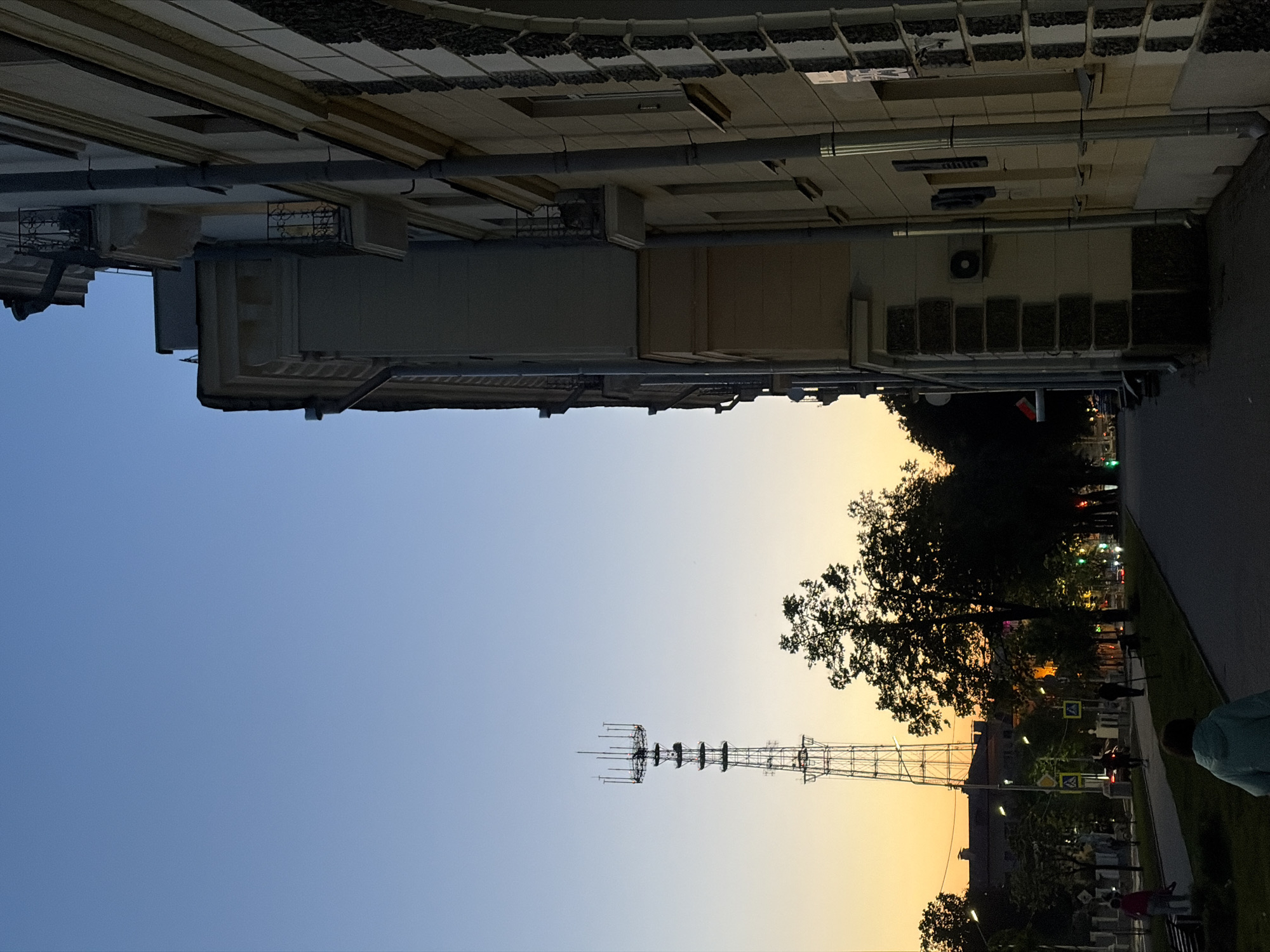 The image shows a street in an unknown city, taken at dusk or dawn. The sky transitions from blue to orange near the horizon. A tall metal transmission tower stands out against the sky. A building runs along the top edge of the frame. There are trees and streetlights along the right side of the image. People can be seen walking on the sidewalk.