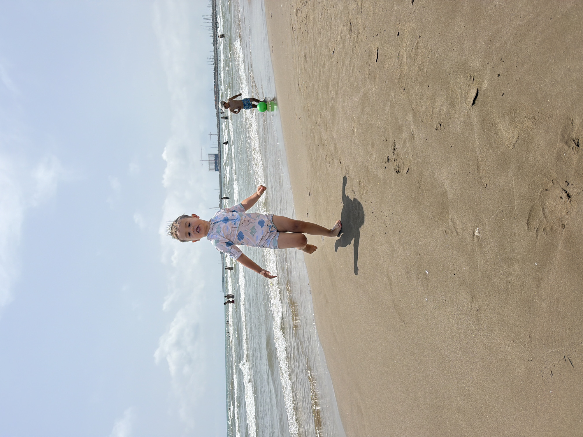 A young child in a light-colored swimsuit with a playful pattern runs towards the camera on a sandy beach. Their right leg is extended forward, mid-stride, and their left arm is bent at the elbow. Behind the child, the ocean stretches to the horizon under a partly cloudy sky. Several other people are visible in the water and on the shore in the distance. Further in the background, construction cranes and sailboats dot the skyline. Another child can be seen playing with a bright green ball near the water's edge. The sand is a light brown, with ripples and footprints indicating recent activity.