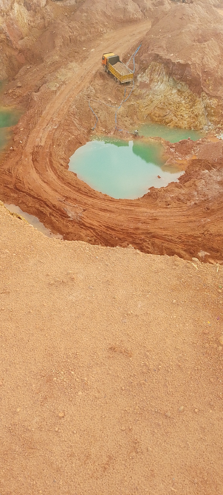 An aerial view shows a large, excavated pit. The pit is characterized by sheer walls of reddish-brown earth, revealing layers of sediment and rock. A turquoise body of water has collected at the bottom of the pit, forming a pool. A bright yellow dump truck with a load of soil is positioned on a dirt path near the edge of the pit. Blue hoses are visible, one extending from the truck and another connected to a pump or water source near the pool. The foreground of the image features a dry, sandy embankment. The scene suggests an active or recently active mining or excavation site.