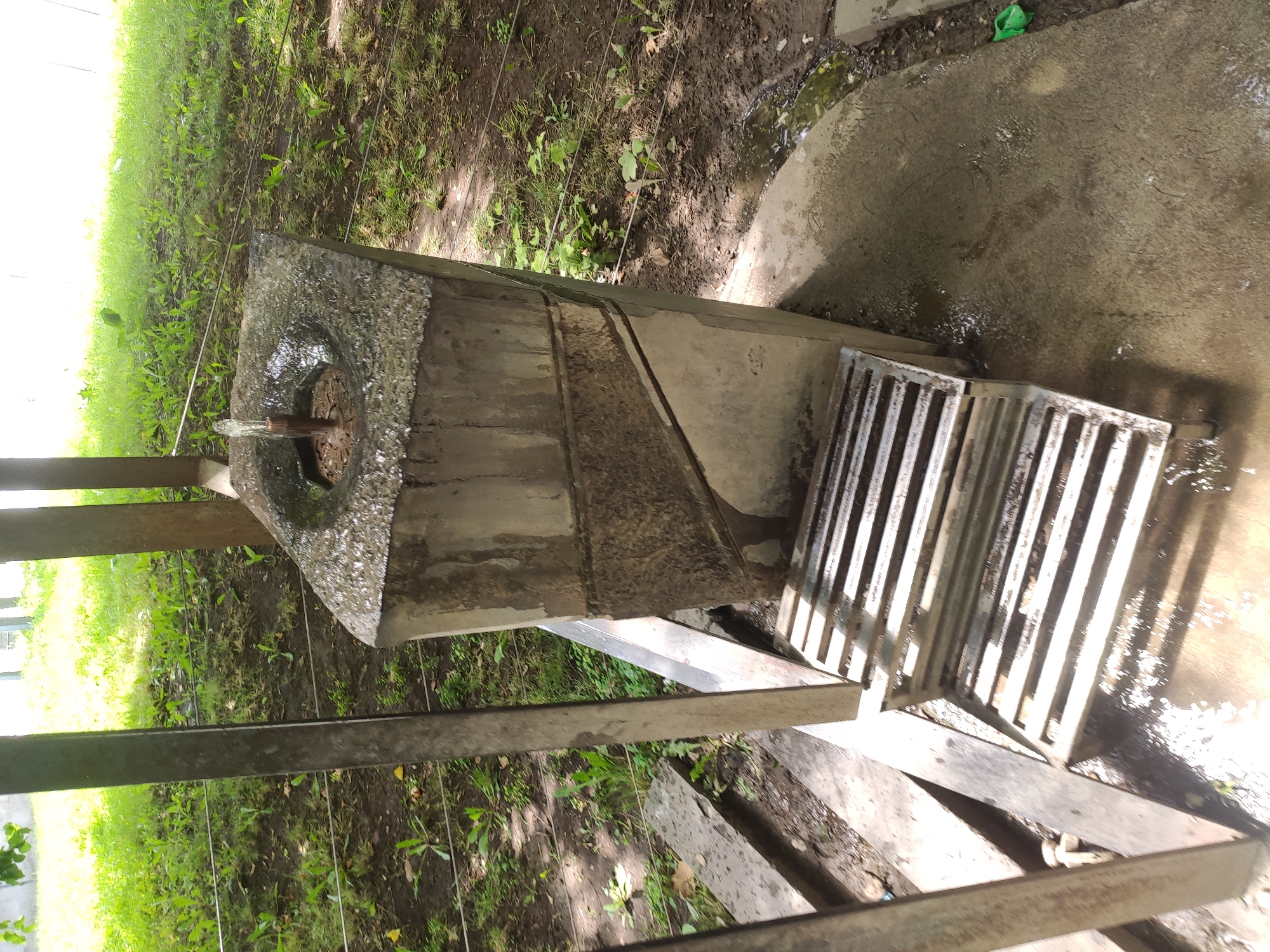 The image depicts an outdoor scene with a concrete fountain that appears weathered and stained. The fountain has a rough, circular opening at the top from which a small stream of water is emerging. The base of the fountain is angular with some textured geometric patterns. In front of the fountain, there is a slatted metal grate, possibly for drainage or to step on. The surrounding environment consists of dirt, grass, and some scattered debris. Several thin, horizontal wires are visible in the background, strung between posts, suggesting some form of barrier or fencing. The overall impression is of a functional but somewhat neglected public amenity. The scene is set in Yerevan, Armenia.