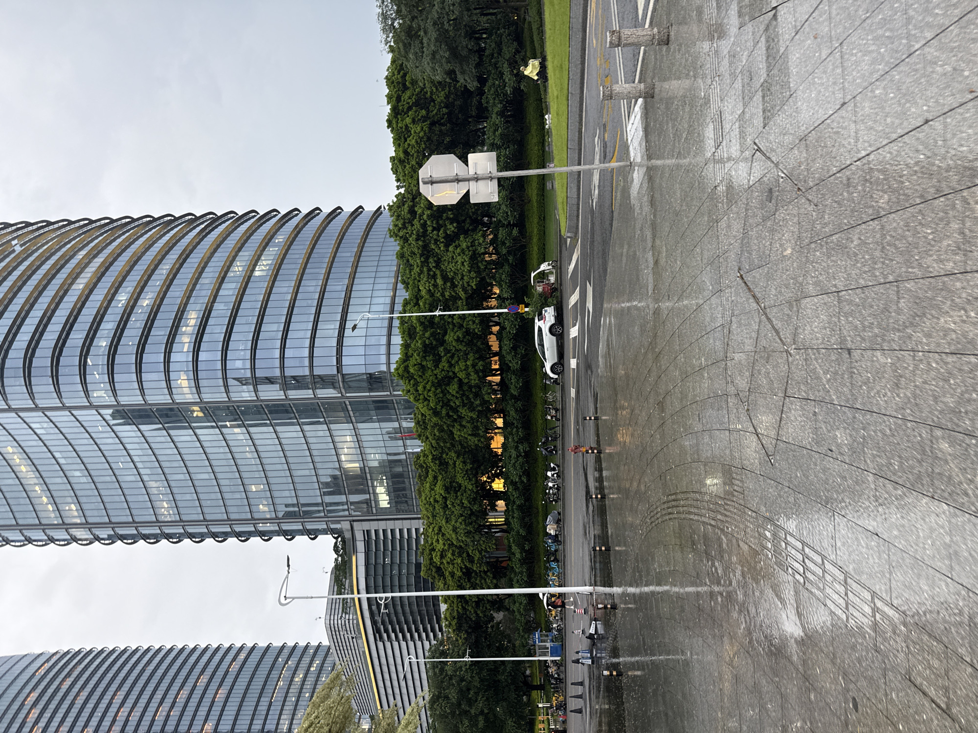 The image depicts a street scene in Haizhu District, China, under overcast skies with wet pavement suggesting recent rain. The foreground is dominated by a wet, patterned stone sidewalk with reflections of the surroundings. To the left, a modern, curved glass-facade skyscraper with visible interior lighting stands prominently. Adjacent to the building, a line of parked motorcycles and electric scooters is visible. Further back, a dense row of mature green trees separates the sidewalk from a road. On the road, a white electric car is parked, and beyond it, a person in a bright yellow jacket is walking. Traffic cones and bollards line the roadside. In the mid-ground, a traffic sign is mounted on a pole. The overall atmosphere is calm and slightly muted, possibly indicating a transition from day to evening or simply a gray day.