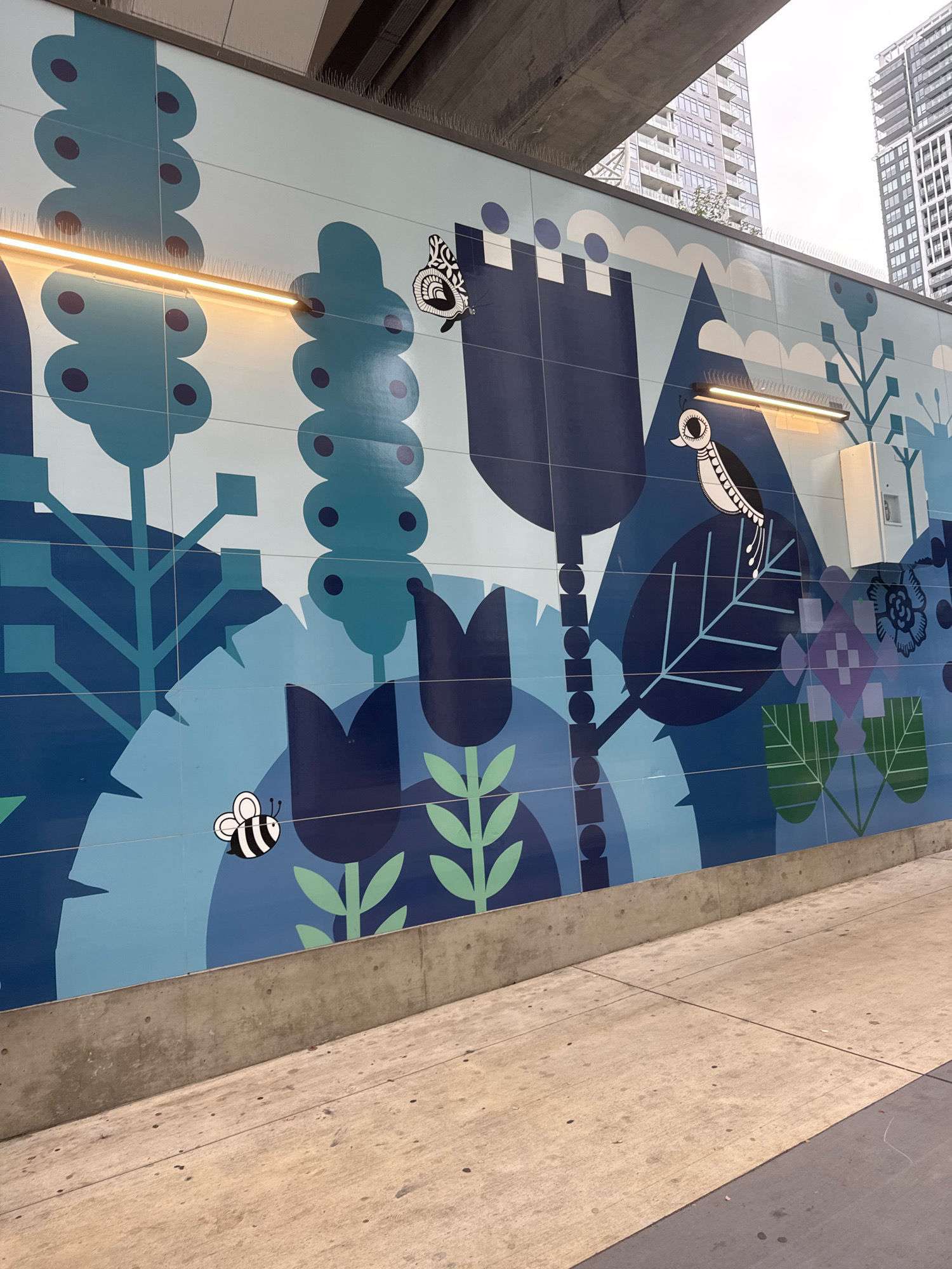 The image shows a mural on a tiled wall in an urban environment, likely a pedestrian walkway or underpass. The mural features a stylized, whimsical depiction of nature with large, abstract plants and flowers in various shades of blue and teal, accented by lighter blue and purple elements. Scattered throughout the mural are small, black and white illustrated insects and birds. A black and white bee is visible in the lower left portion of the mural, and a stylized bird is perched on a large leaf in the upper right quadrant. A butterfly is also present near the top left.
Two industrial-style lights run horizontally across the mural, illuminating the artwork. The wall itself is constructed of rectangular tiles, with visible grout lines. At the base of the wall is a concrete barrier, and in front of this is a concrete walkway, which appears to be gently sloping upwards and to the right. In the background, beyond the mural, are tall modern buildings, suggesting an urban setting. The sky is overcast.
This mural is located in Burnaby, Canada, and is part of the outdoor art installations at the Brentwood Town Centre SkyTrain station. The installation is designed to add visual interest and a sense of nature to the urban infrastructure.