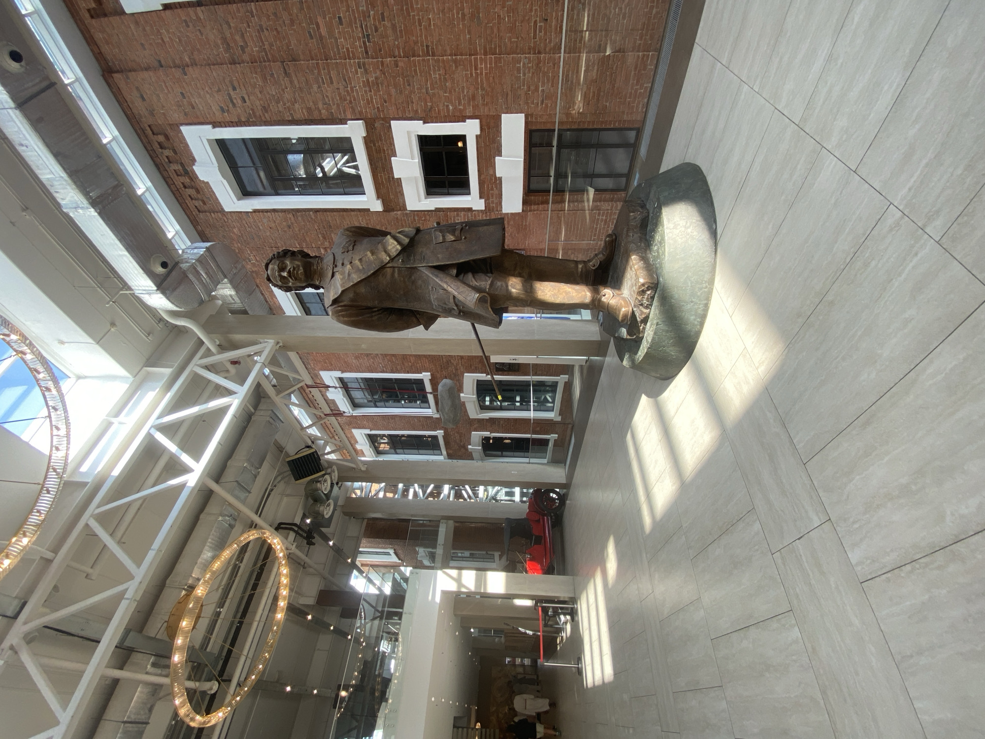 The image depicts a bronze statue of Vasily Tatishchev standing in the main atrium of the Yeltsin Center in Yekaterinburg, Russia. Tatischev is standing on a green stone pedestal. The building's interior architecture is visible with white support beams and large windows showcasing the building's brick exterior. Overhead lighting fixtures of large hoops are visible. The floor is paved with light-colored stone tiles. Red car is seen in background. People are seen walking the hall.