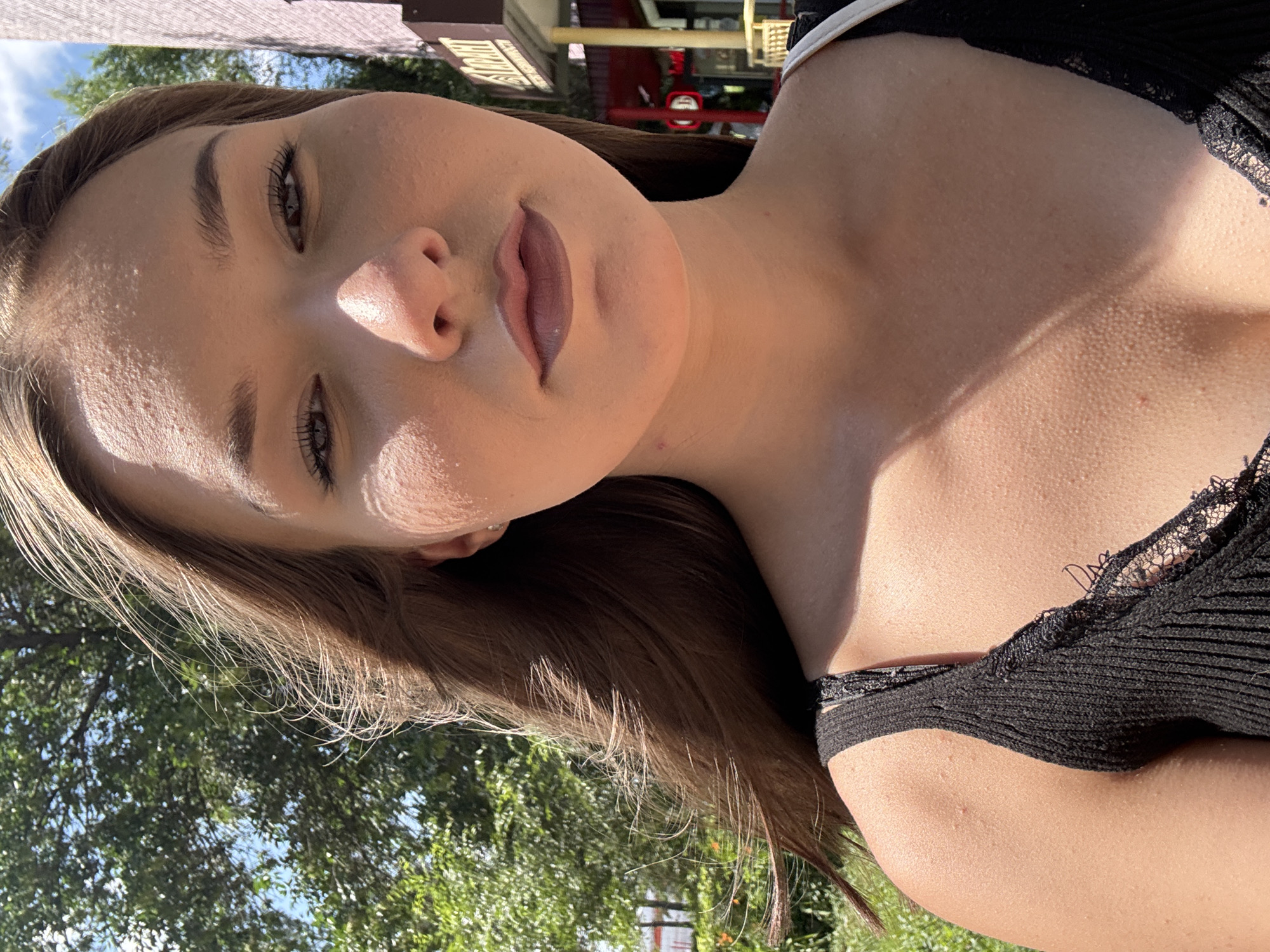 In a close-up, outdoor selfie, a young woman with long, brown hair is pictured. She is wearing a black tank top with lace detailing. Her hair is blown to the side, revealing her face, which is illuminated by natural sunlight. Her makeup is subtle, with defined eyebrows and lipstick. Her skin appears to have some texture, possibly from minor blemishes or pores. The background shows a blur of green foliage, suggesting a park or tree-lined street, and a hint of a building with signage. The overall impression is a casual, sunlit moment captured by the subject herself. The city of Belovo, Russia is not identifiable from the visual cues present in the image.