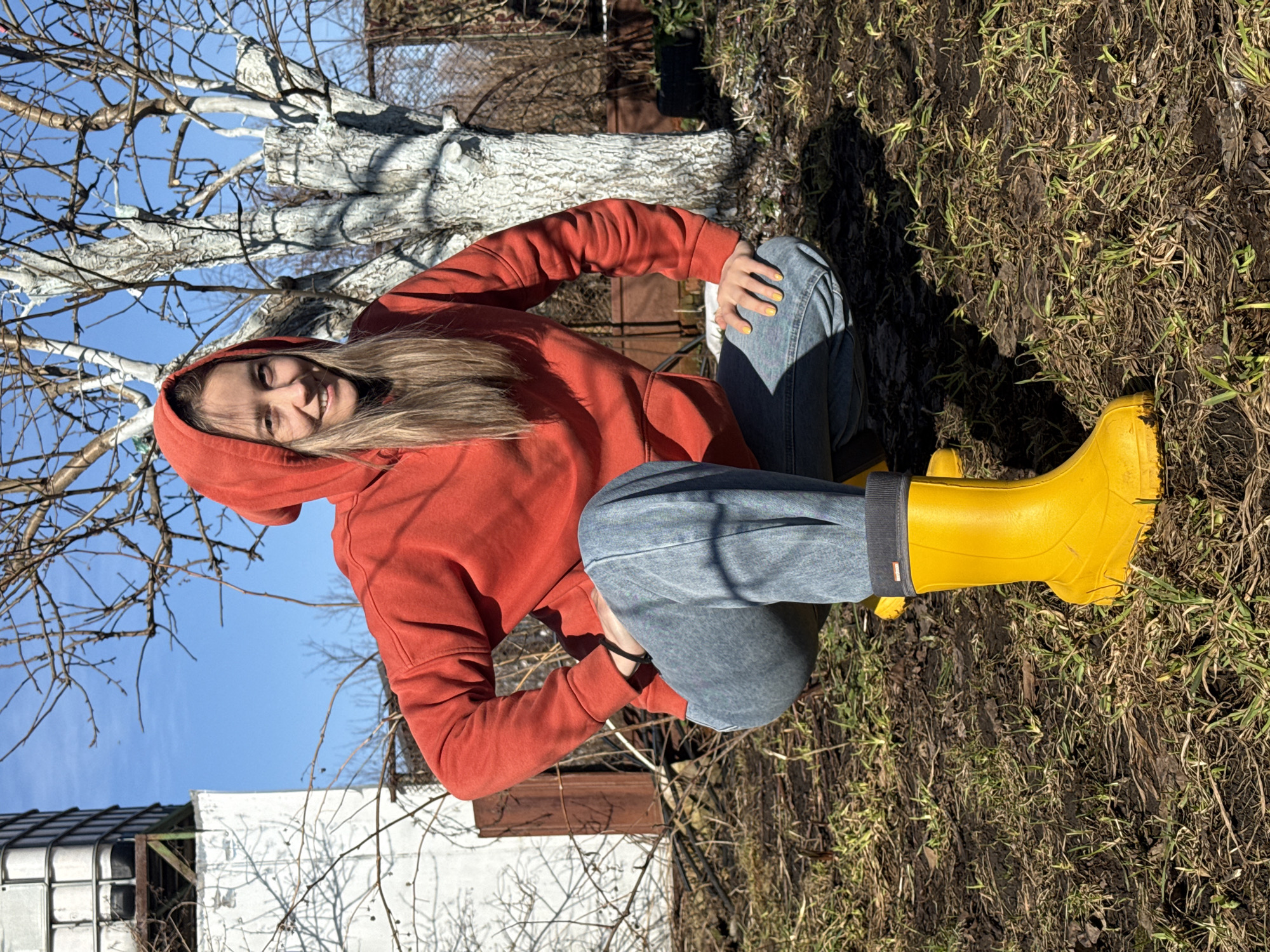 A young woman is kneeling outdoors on soil covered with sparse grass, wearing an orange hooded sweatshirt, blue jeans, and bright yellow rubber boots. She is smiling and looking at the camera. Behind her is a tree with white-painted branches and a clear blue sky. In the background, a white building and a brown fence are partially visible. The image appears to be taken on a sunny day. The location is Chaikovsky, Russia.