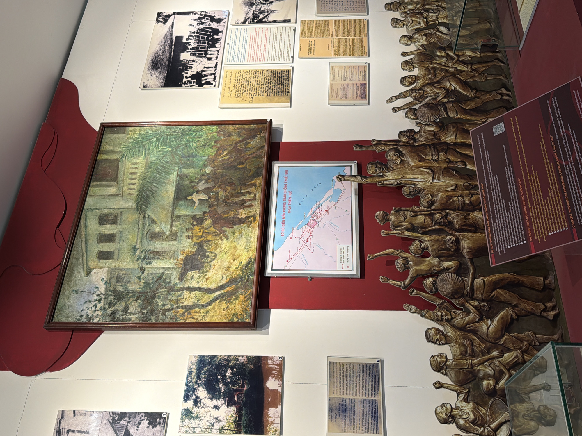 The image depicts a museum exhibit dedicated to a historical event, likely related to Vietnamese protests or resistance. In the background, a large, framed painting shows a building with a crowd of people, some on foot and some with a cart, in a verdant setting. To the right of the painting are framed black and white photographs, one showing a group of people in formal attire, and another, further right, of a historical scene with soldiers. Below the main painting, a map labeled "SỞ ĐỒ BIÊN PHÒNG THUÀN THIÊN 1908" (Map of Thien Thua Border Defense 1908) is displayed. Scattered on the wall around these items are several smaller framed documents and photographs, some appearing to be historical texts or proclamations.
The foreground is dominated by a large, bronze-colored bas-relief sculpture depicting a dynamic crowd of figures in various poses, many with their arms raised in a gesture of protest or celebration. The figures are rendered in a stylized, almost abstract manner, conveying a sense of movement and collective action. Below the sculpture, a red display board with text in both Vietnamese and English provides information about "THE SCENE: HUE'S MOVEMENT OF PROTESTATION CONTRE LES IMPÔTS À HUE EN 1908" (The Scene: Hue's Movement of Protestation Against Taxes in Hue in 1908). The text details the historical significance of the event, which occurred in April 1908, and its impact. The environment appears to be an indoor exhibition space with white walls and a red accent wall or feature behind the central painting and map. A glass display case is visible in the lower left corner, reflecting some of the sculpture. The overall impression is one of historical commemoration and educational display.