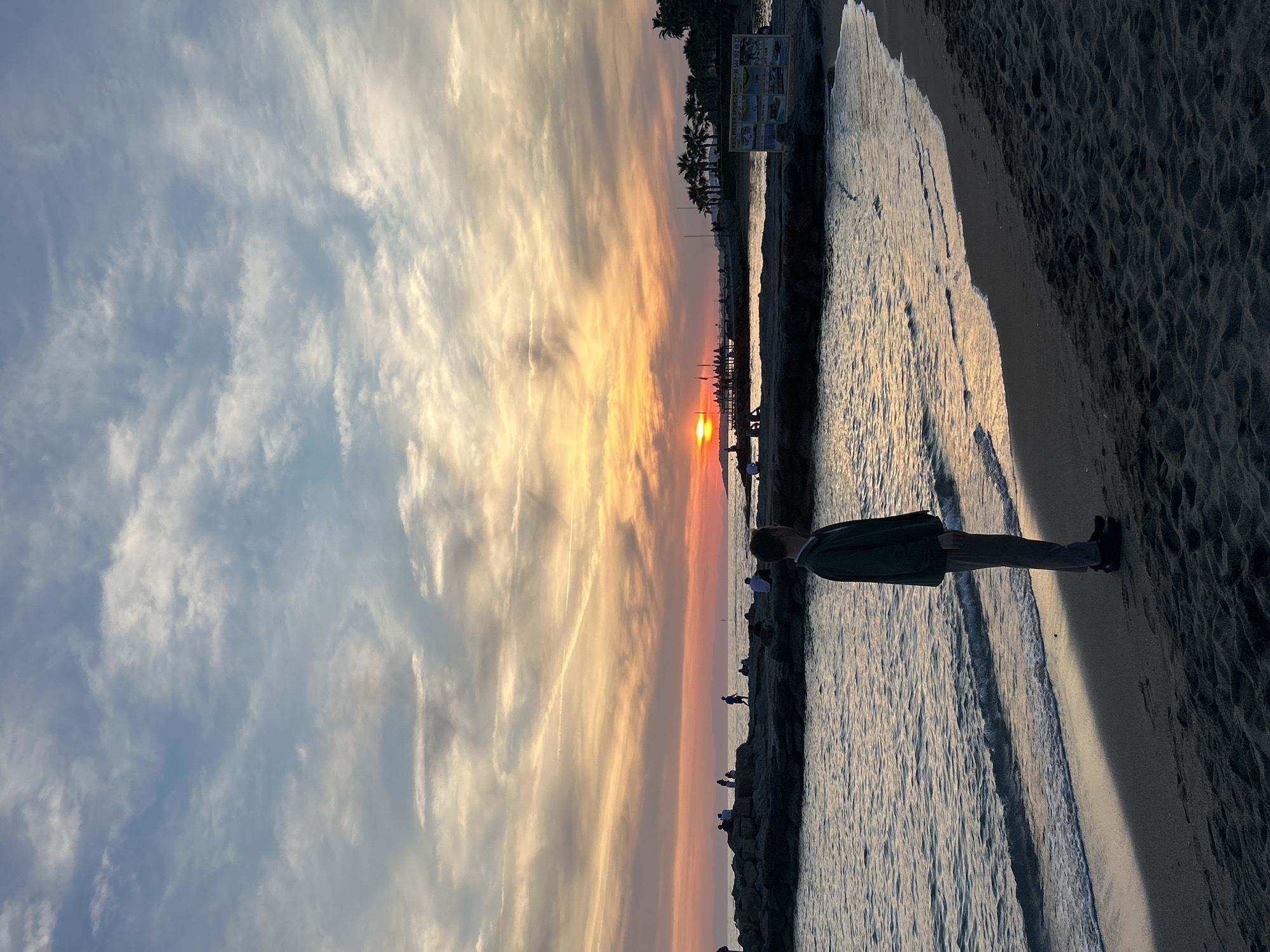 The image captures a sunset scene in Alanya, Turkey. A person stands on a sandy beach beside the ocean. The setting sun casts a warm glow on the water, the shoreline, and a bank of clouds. In the distance, a breakwater with people standing on it is visible. Further, there are silhouettes of palm trees and buildings on the coastline.