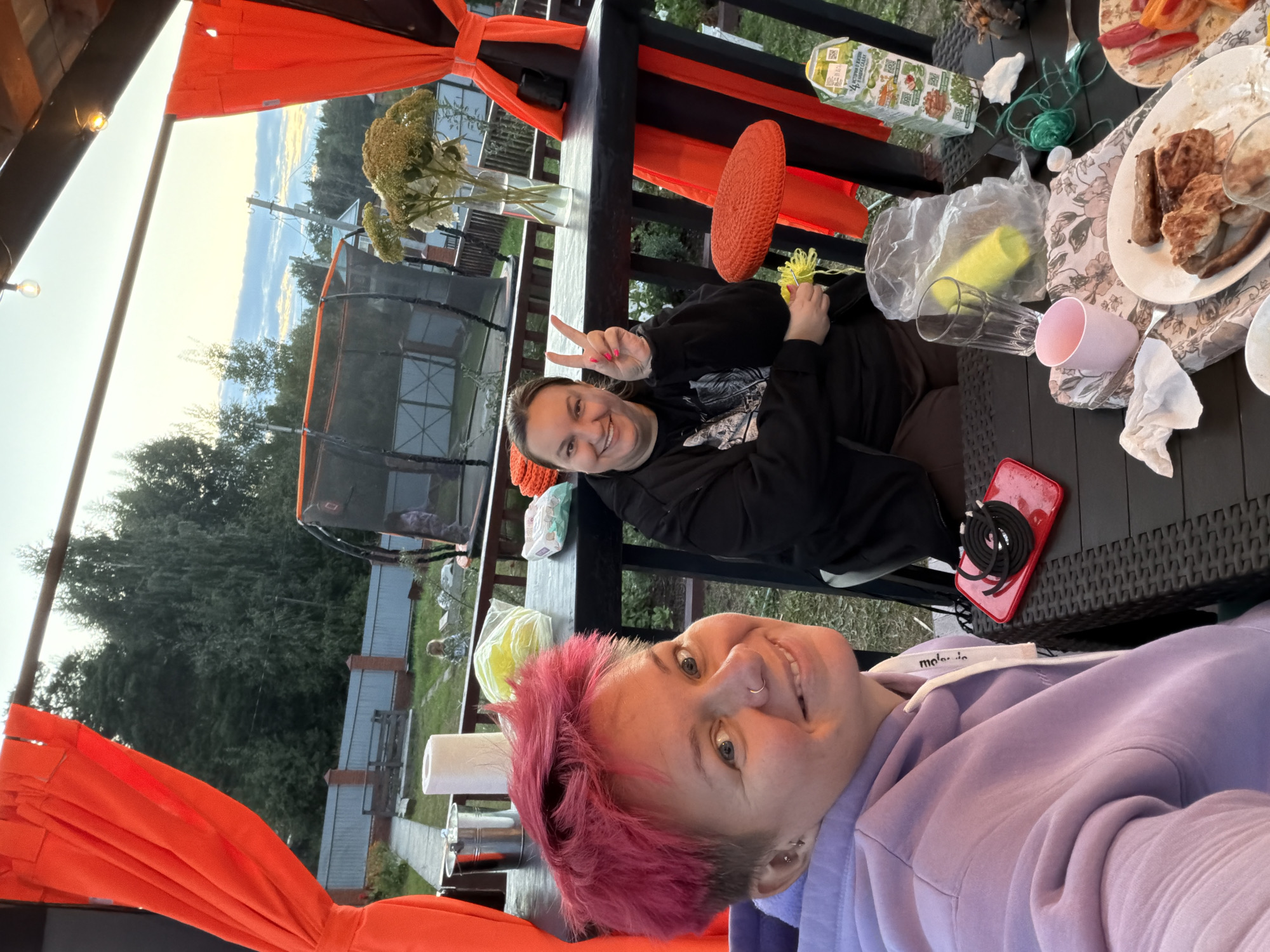 Two women are seated at a table under a covered outdoor structure, likely a gazebo or patio, with orange curtains visible. The woman on the left has short, bright pink hair, a nose ring, and is wearing a light purple hoodie. She is smiling and looking at the camera. The woman on the right has dark hair and is wearing a black hoodie. She is smiling and holding up two fingers in a peace sign. On the table, there are plates with food, drinks, cups, and other miscellaneous items. In the background, a trampoline and trees are visible, suggesting a residential or outdoor recreational area. The sky appears overcast with some lighter clouds. The location is identified as the city of Chaikovsky, Russia.
