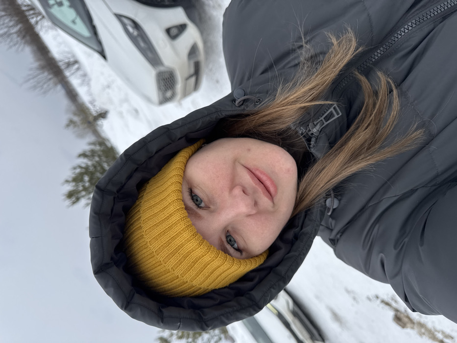 The scene is set in Zaosinovo, Russia, outdoors during winter conditions. A female subject is positioned in a close-up, direct-to-camera view, appearing to take a selfie. She has blue eyes, light brown hair, and is wearing a bright yellow ribbed knit beanie under a dark grey/charcoal puffer jacket with the hood up, framing her face. A faint smile is present on her lips. The background consists of snow-covered ground and distant, sparsely treed hills under an overcast sky. A white SUV, possibly a Kia, is partially visible to the right, parked on the snowy terrain. Another light-colored vehicle is partially visible to the far left.