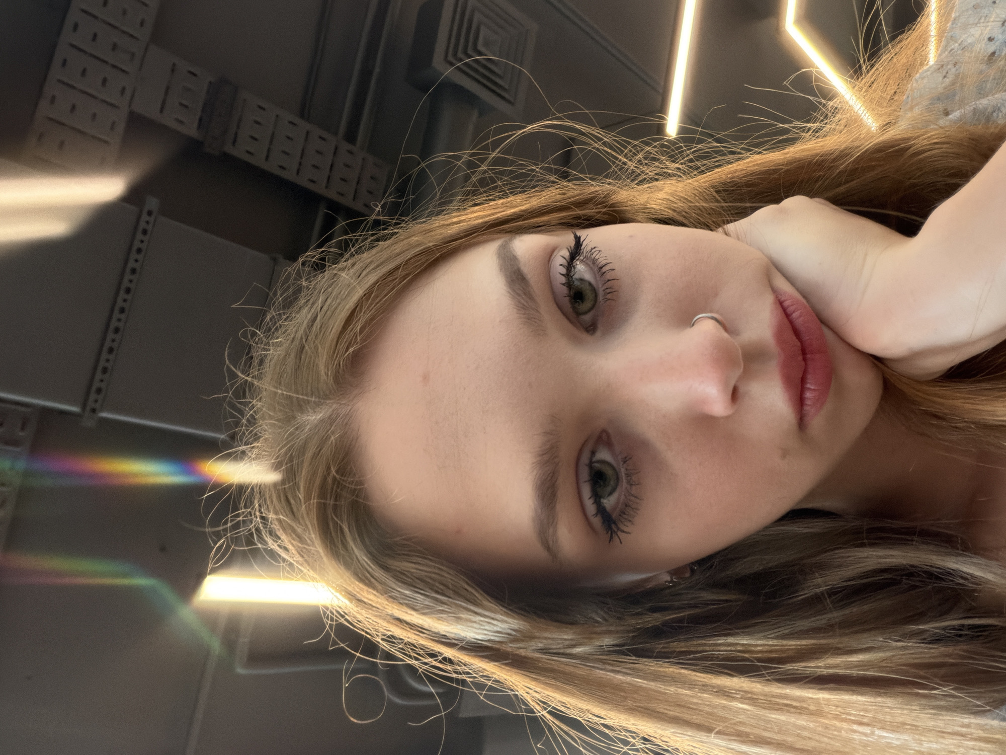 A young woman with long, blonde hair, a nose ring, and light makeup is leaning on her hand and looking down at the camera. A rainbow light refraction is visible in the upper left corner of the image, reflecting off a ceiling with visible ventilation ducts and light fixtures. The environment appears to be indoors, possibly an office or public building.