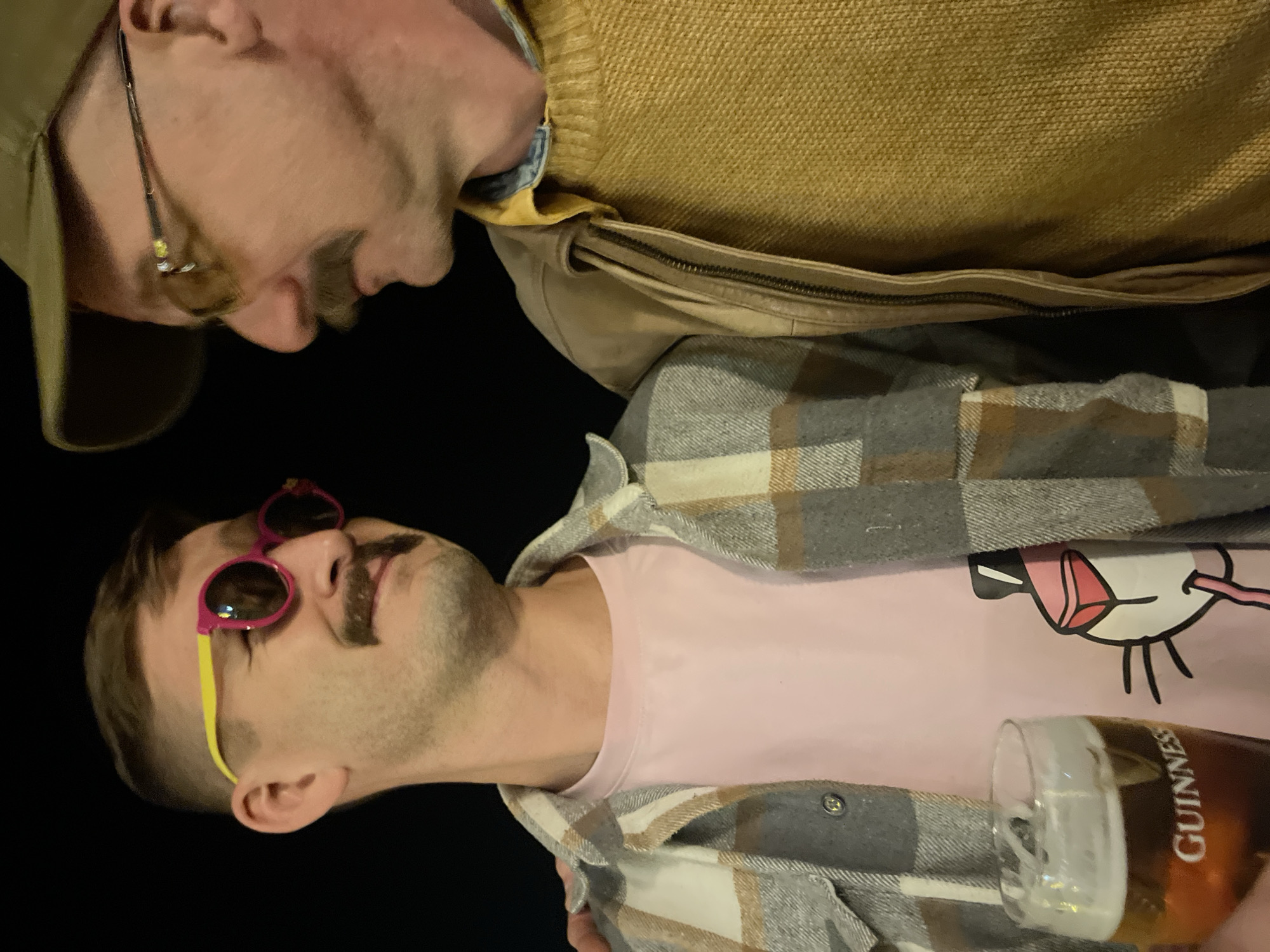 Two men are closely positioned. One man wears a brown hat, glasses, a brown sweater, and a tan jacket. The other man wears pink and yellow sunglasses, a pink t-shirt with a cartoon design, and a gray and tan plaid flannel shirt. He holds a pint of Guinness beer. The location is not identifiable based solely on this image.