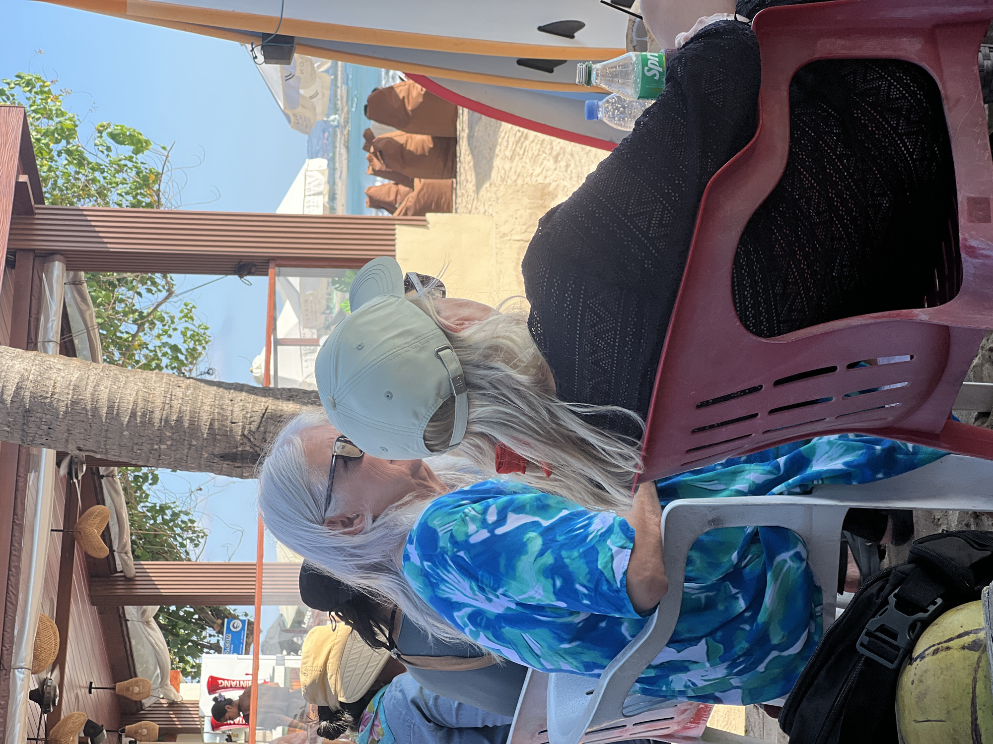 The image captures a scene at a beachside establishment in Kuta, Indonesia, likely a restaurant or cafe, with a view of the ocean. In the foreground, two women are seated. The woman on the left has long, gray hair and is wearing glasses and a blue and green tie-dye shirt. The woman on the right is wearing a light green baseball cap and a black, lace-patterned top. Both are facing away from the camera, looking towards the ocean.
The environment features elements of a tropical beach setting. A thatched roof structure with hanging lamps provides shade overhead. A large tree trunk, likely a palm tree, is prominent on the left side of the frame. In the background, the sandy beach transitions to the blue ocean, with a few boats visible in the distance. Several large, brown beanbag chairs are arranged on the sand. A white umbrella is also visible. Surfboards stand upright on the right side of the image, indicating the location's proximity to surfing activities. On a table in front of the seated women are two bottles, one appearing to be water and the other a bottle of Sprite. A black backpack and a yellow, round object, possibly a coconut, are on the ground near the woman on the left. The overall atmosphere suggests a relaxed, daytime setting at a popular beach destination.