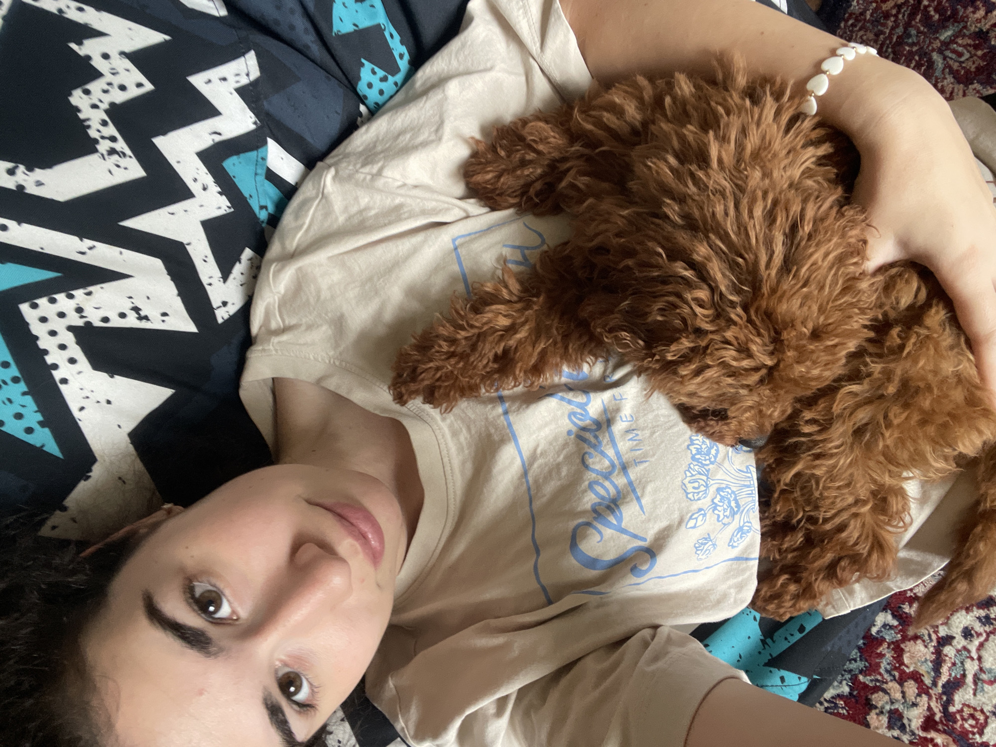 A woman with dark hair and brown eyes lies on a bed. She is wearing a light-colored t-shirt with a blue graphic on the front. A small, brown, poodle-like dog is lying on her chest. She is holding the dog with one arm. The bed is covered with a geometric patterned blanket in black, white, and turquoise. The woman is looking directly at the camera with a slight smile. Part of a patterned rug is visible in the lower right corner. There is no indication in the image that this is in Sofia, Bulgaria.