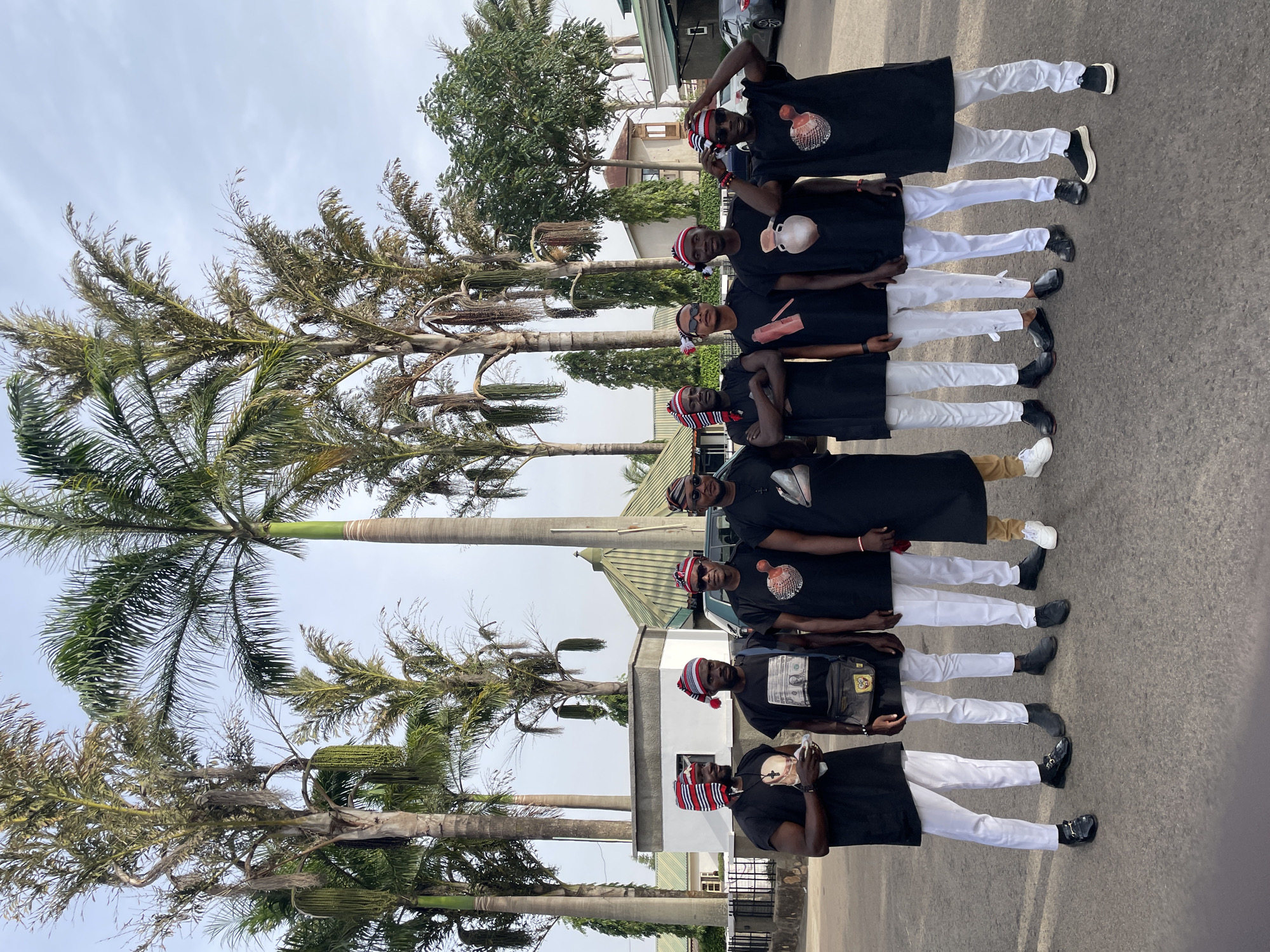A vertical image shows six men in a line along a street.
The men all wear black shirts and white pants. Most wear striped hats. The location appears to be a city in Unknown, Unknown, indicated by the buildings and palm trees. The street is asphalt.