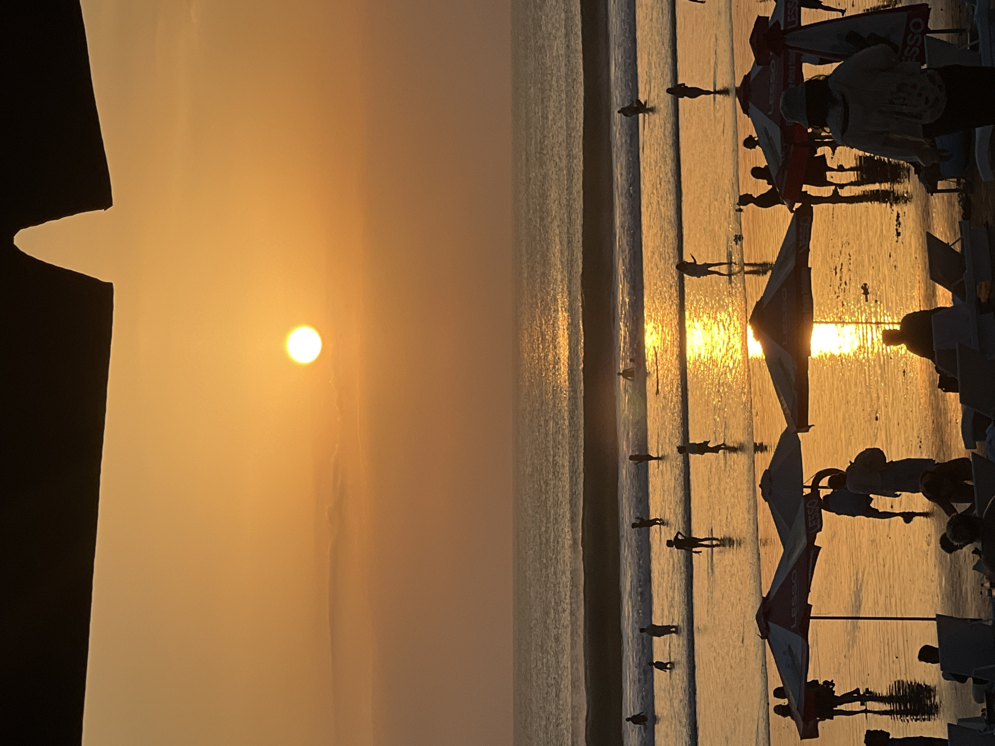 The image depicts a sunset scene at Kuta Beach, Indonesia. Silhouettes of people are visible on the beach and in the shallow water, engaged in various activities. Some individuals are standing, while others appear to be wading or walking. Umbrellas are set up on the sand, with a few people gathered beneath them. The ocean has gentle waves, and the setting sun casts a warm, golden glow across the water and the wet sand, creating reflections. The sky is a gradient of orange and yellow, with the sun a bright, distinct circle. The overall atmosphere is serene and typical of a beach at dusk.