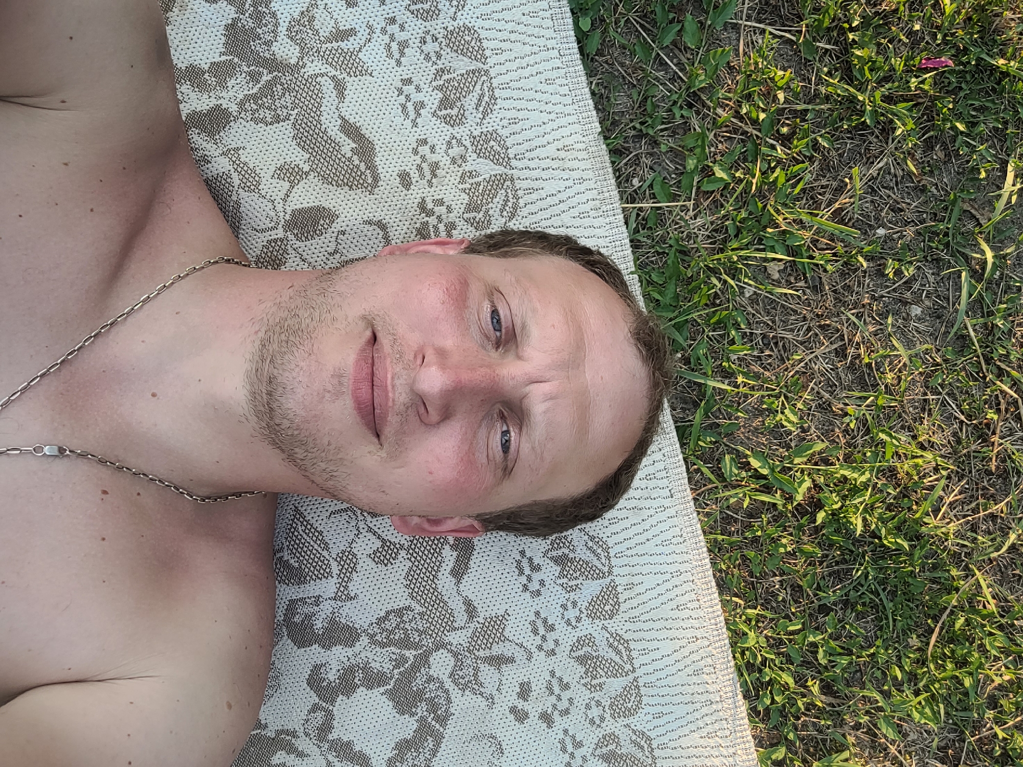 A man with fair skin, light brown hair, and blue eyes is lying on his back on a blanket outdoors. He is shirtless and wearing a silver chain necklace. The blanket has a patterned design in beige and brown. Behind the blanket is a patch of grass and dry dirt, with some green weeds growing. The lighting suggests it is daytime. The man's head is tilted back, and he is looking directly at the camera with a neutral expression. His skin appears slightly flushed, possibly due to sun exposure. The image is a close-up, focusing primarily on the man's face and upper torso.