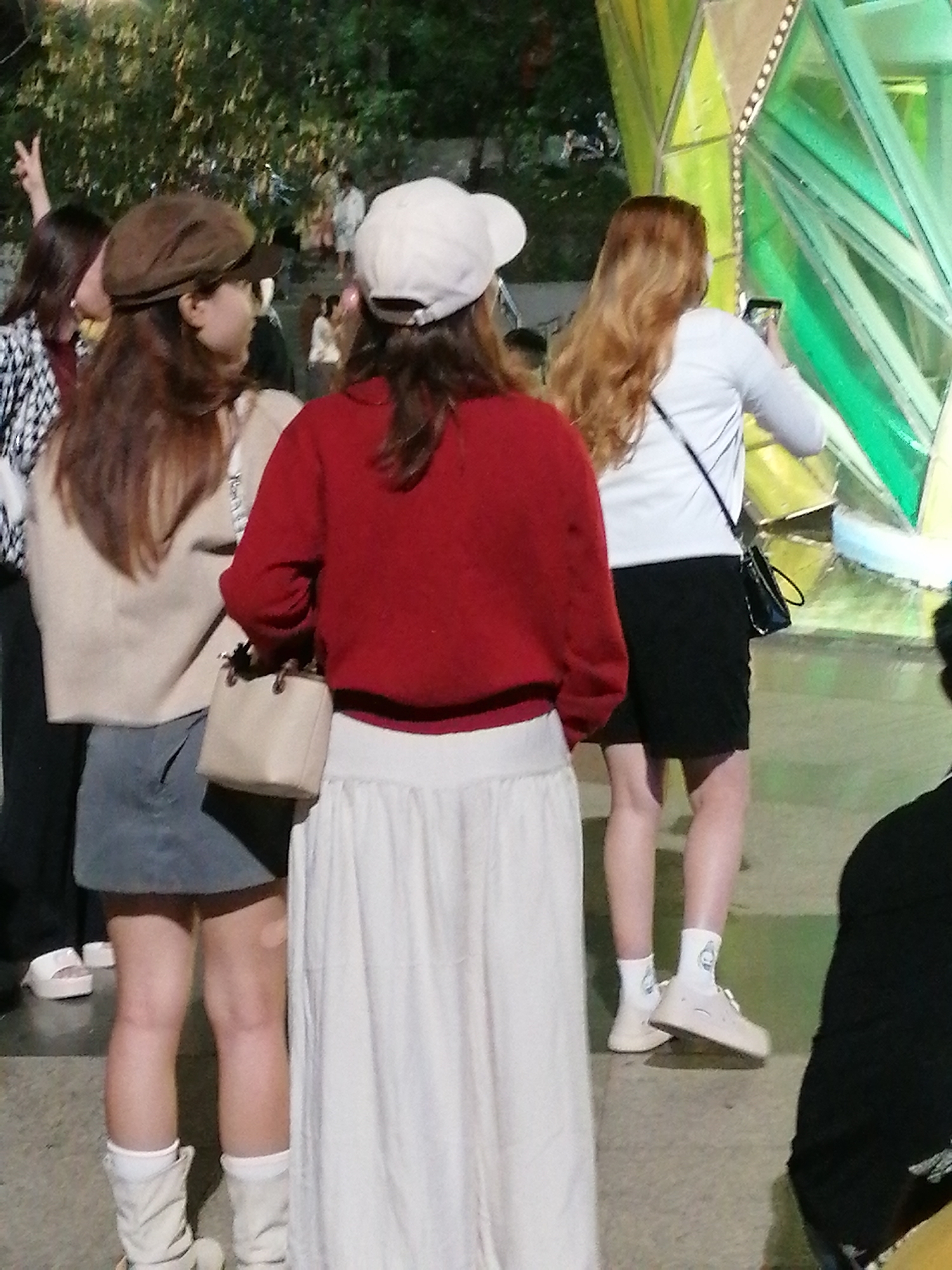 Three women are standing outside near a large, colorful, geometric structure. The woman on the left is wearing a patterned shirt, a beige jacket, and a grey skirt, with white boots. The woman in the middle is wearing a red sweater, a white skirt, and white boots. The woman on the right is wearing a white long-sleeved shirt and a black skirt, with white socks and sneakers. The woman in the middle is holding a beige purse. The woman on the right is holding a phone and appears to be taking a picture. The scene is lit with artificial lights, suggesting it is evening or night. The overall atmosphere appears casual, possibly at an outdoor event or festival. The location context is Da Lat, Vietnam.