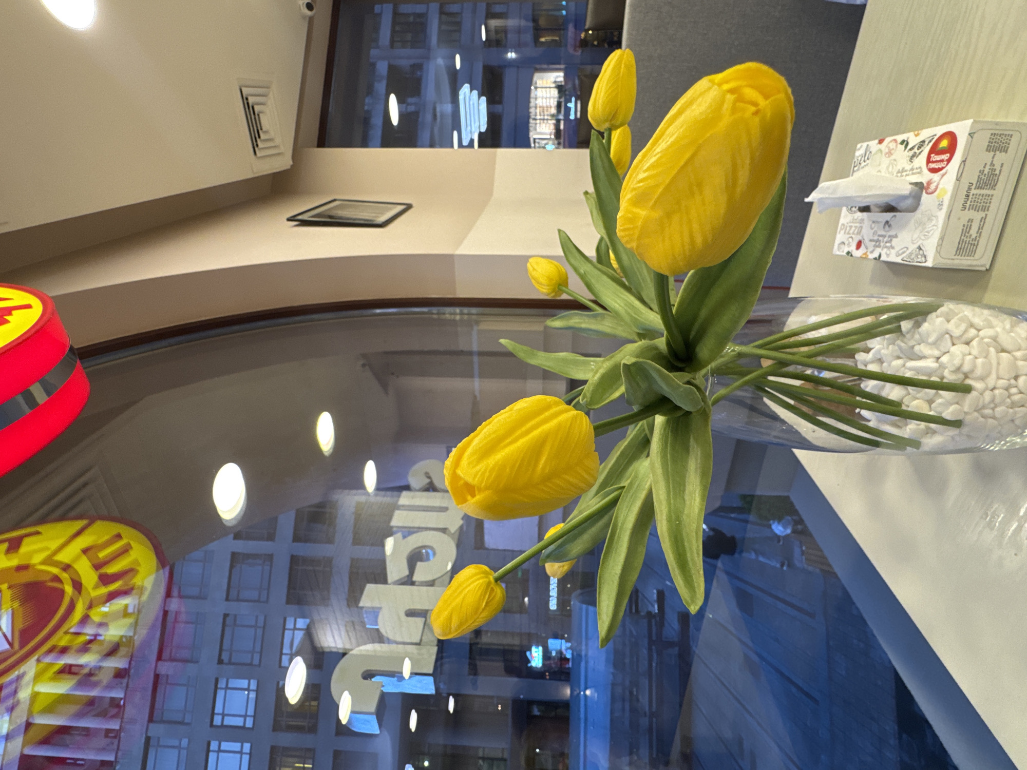 The image shows a vase with yellow tulip flowers placed on a table next to a tissue box. A window behind the flowers offers a reflected view of a city street with signage. The location is Yerevan, Armenia, as indicated by the Armenian text on the tissue box.