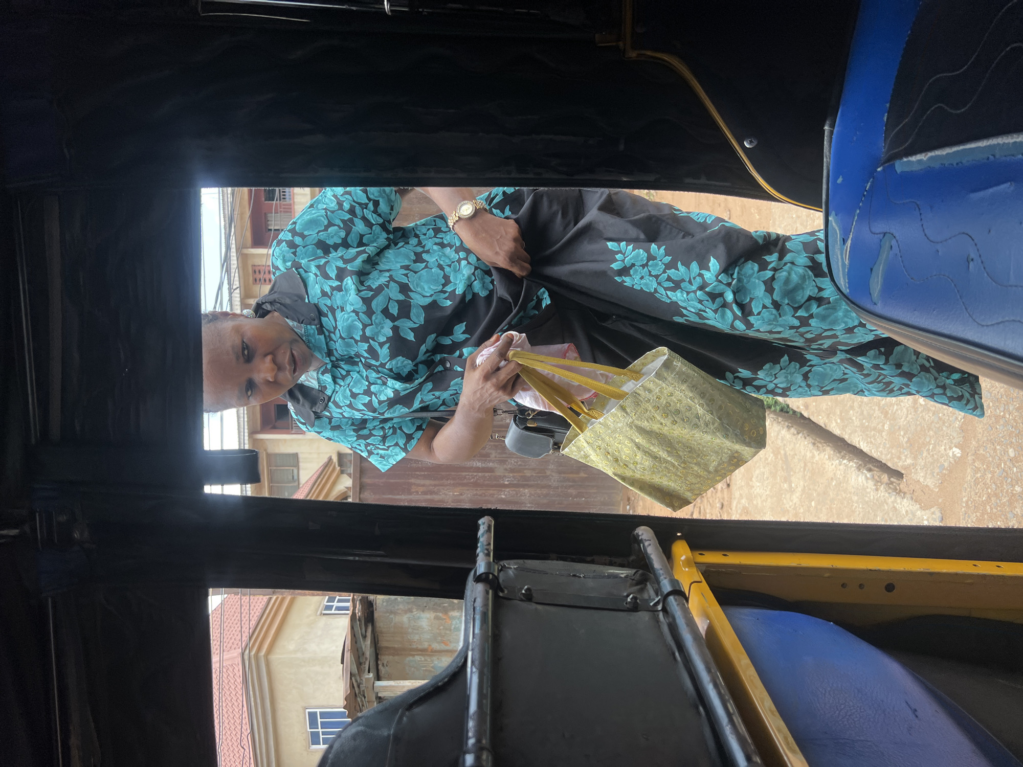 Captured from inside a vehicle, the photograph presents a woman holding a gold-colored bag. She is adorned in a turquoise and black patterned dress and a gold wristwatch is visible on her wrist. The vehicle's interior, featuring black and blue seats, is visible, framing the scene. Outside the vehicle, buildings line the street, hinting at an urban setting. The woman is positioned in such a way that she seems to be looking into the vehicle.