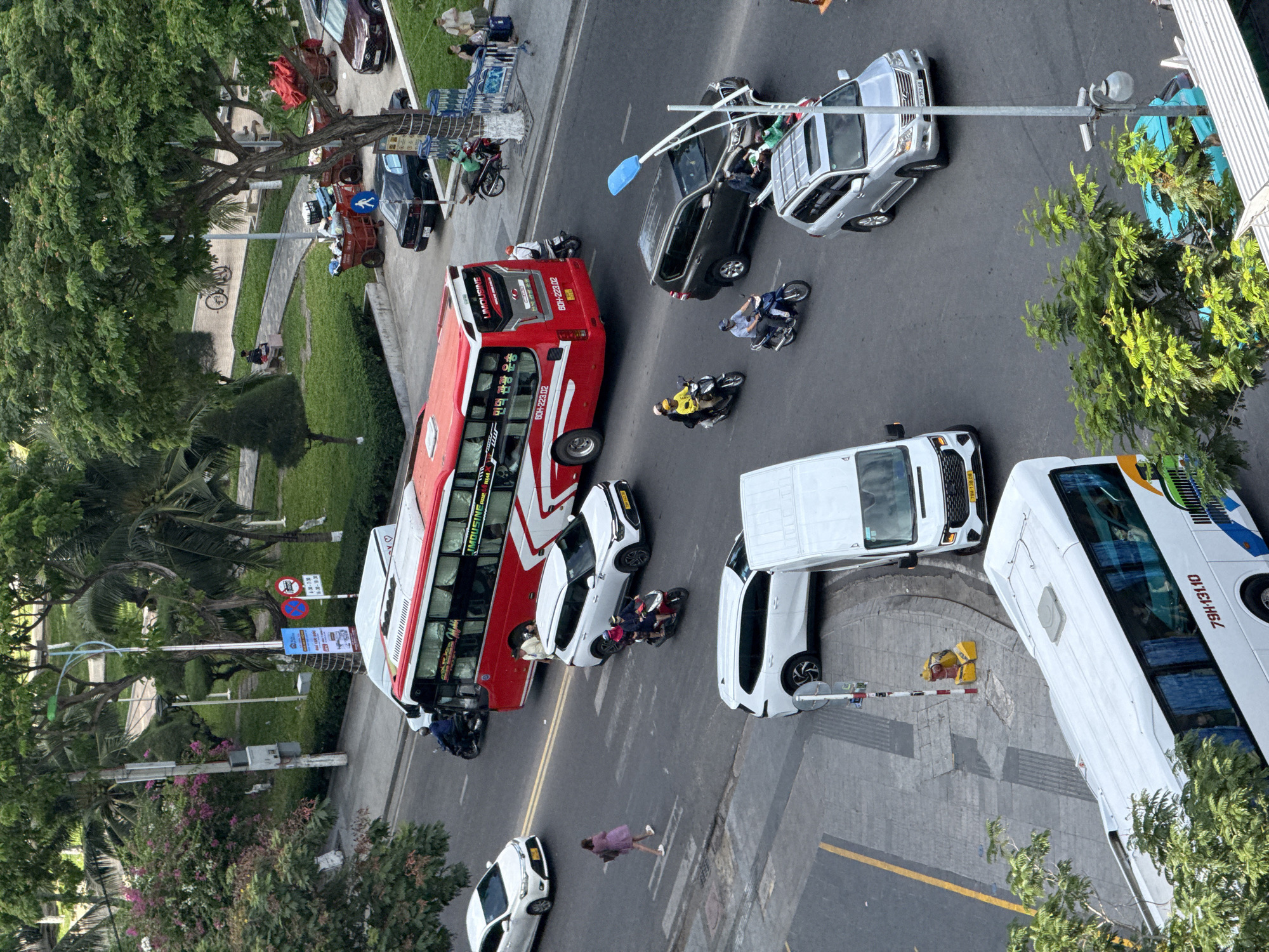 In Nha Trang, Vietnam, an aerial view captures a busy street scene. A large red and white tour bus is a prominent feature, its side displaying Korean text and the license plate "60H-223.02." Several vehicles, including white cars, a dark SUV, a silver SUV, and a white van, navigate the asphalt road. A woman in a pink dress is seen crossing the street. Multiple motorcycles are also present, some carrying passengers. The street is bordered by a park area with lush green trees, grassy lawns, and paved walkways. Parked carts filled with goods are visible in the park. A person is seated on a bench in the park, and another person is standing near a building entrance in the background. Lampposts and traffic signs are also part of the urban landscape.
