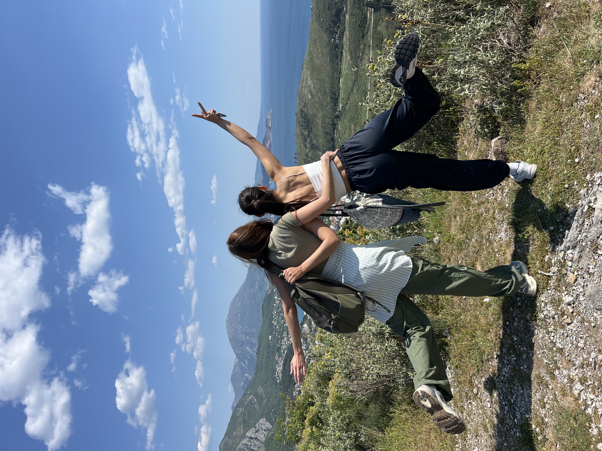 Two young women stand on a grassy hillside overlooking a mountainous coastal landscape under a blue sky dotted with white clouds. One woman wears a white tank top and black sweatpants, standing on the edge with her right arm raised in a peace sign. The second woman, wearing a green t-shirt, striped shirt, and green pants, stands behind her, embracing her. A backpack is slung over her shoulder. The scene is shot from a slightly elevated perspective, with the landscape featuring hills, valleys, and the Adriatic Sea in the distance. The location is Zagrađe, Montenegro.