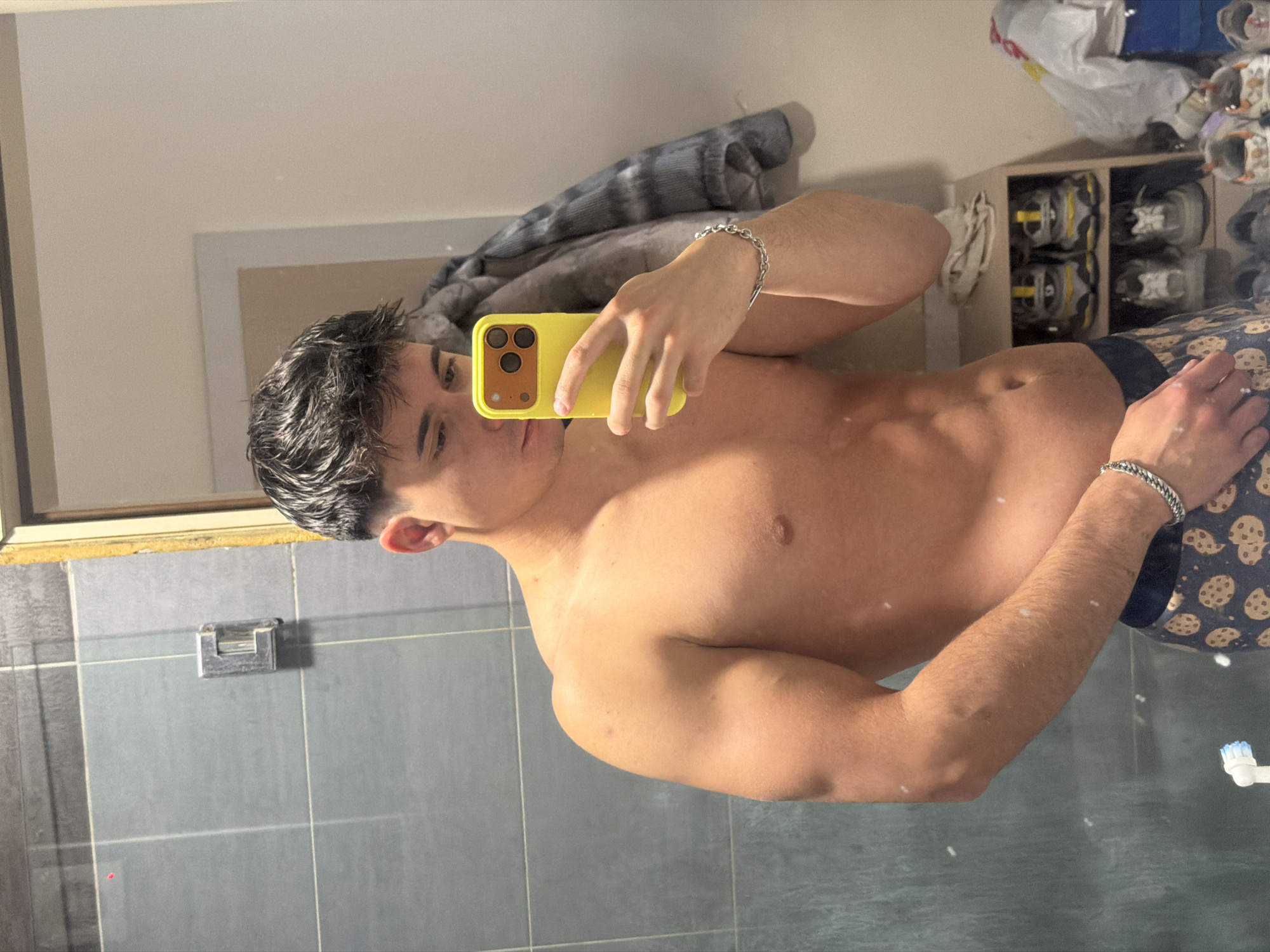 A young man is taking a selfie in what appears to be a bathroom. He is shirtless and wearing blue shorts with cookie patterns. He is holding a yellow phone up to his face, obscuring most of it. The background includes a bathroom mirror reflecting the man and part of the bathroom. There is a towel or clothing item draped over a shelf-like structure behind him, and a shelf with shoes. The bathroom has gray tiled walls. The location is not discernable from the image and cannot be said to be located in Sofia, Bulgaria.