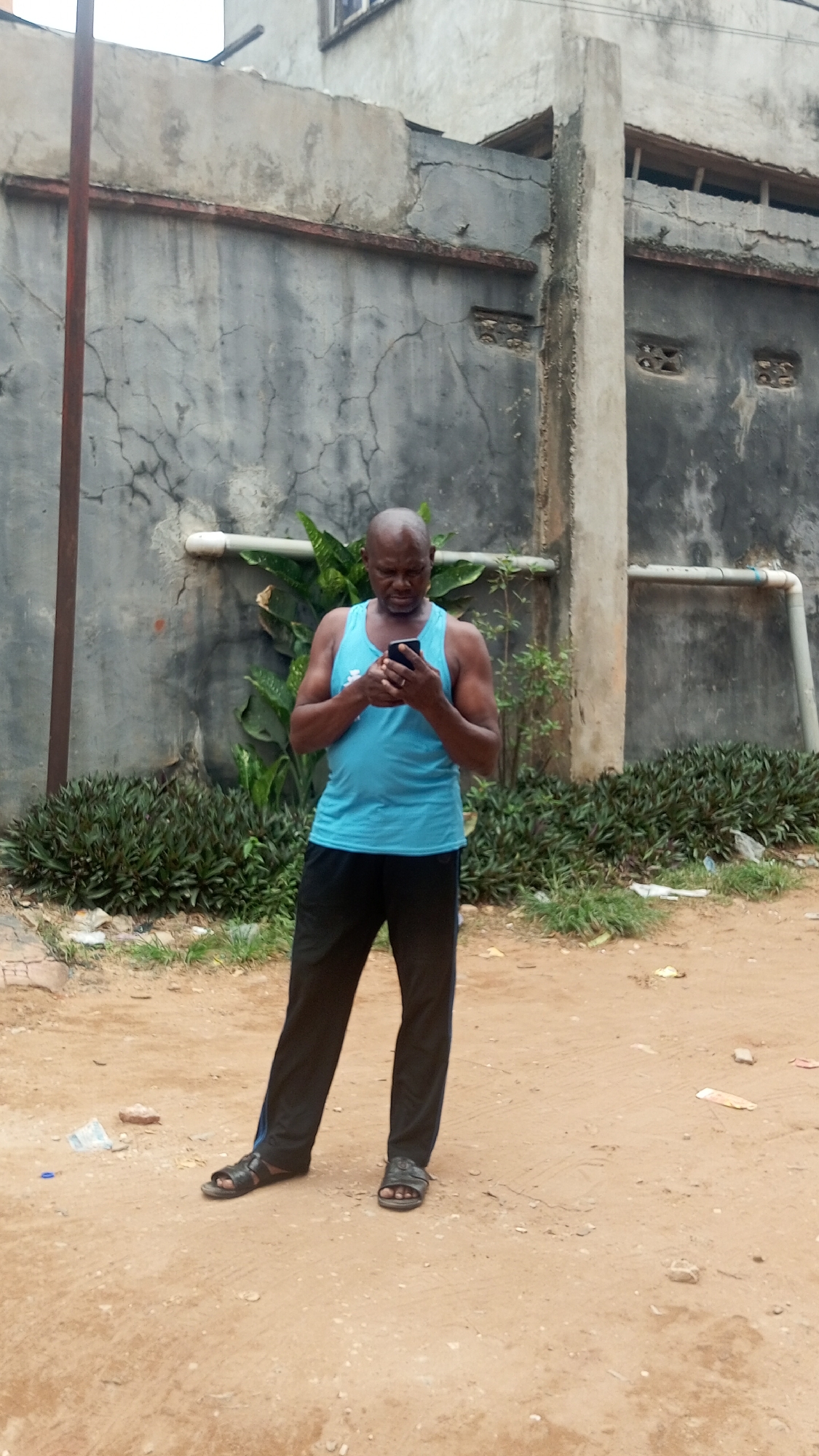 A middle-aged Black man stands on a dirt surface, facing forward and looking down at a smartphone in his hands. He is wearing a light blue tank top, dark-colored pants, and sandals. Behind him is a concrete wall with visible cracks and discoloration, as well as greenery planted along its base. A pipe is attached to the wall. The background includes a section of a building with windows. Debris litters the dirt ground. The location is Obosi, Nigeria.