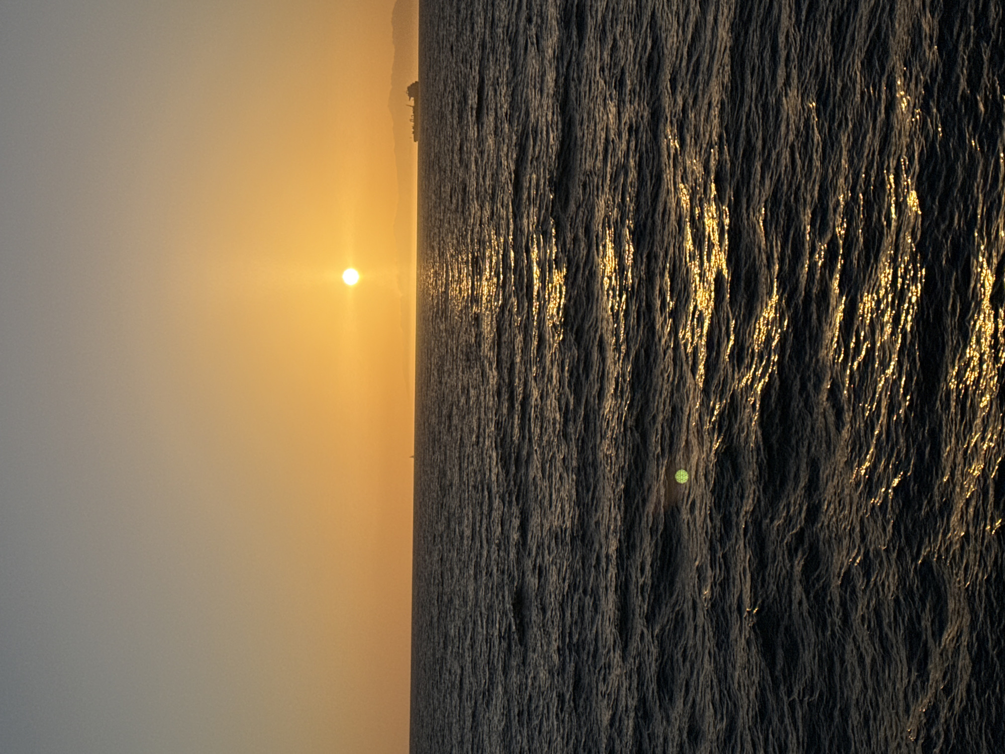 A vertical, wide shot captures the sun setting over the ocean in Manhattan Beach, California. The sky is a gradient of soft yellow and pale blue. The sun, a bright orb, is positioned slightly to the left of the frame, casting a shimmering golden path across the dark, choppy water. In the distance, a silhouette of a ship and a faint outline of hills are visible on the horizon. A single sailboat is also visible in the far distance to the left. A subtle lens flare is present in the lower left quadrant of the image.