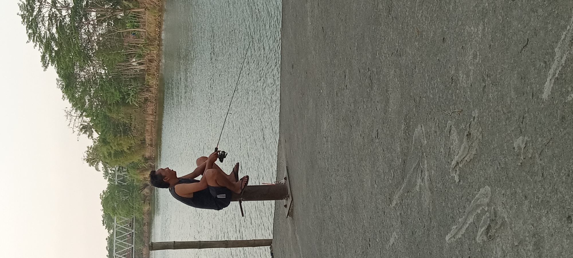 An Asian man is fishing from a concrete pier at the edge of a body of water, likely a river. He is crouched on a metal pole that is bolted to the concrete. The man is wearing a dark sleeveless shirt and dark shorts with white stripes. He is holding a fishing rod and reel. In the background, a white metal bridge with a lattice design crosses the water. The far bank of the water is lined with lush green trees and vegetation. The sky is overcast and pale. The concrete pier has visible tracks or marks on its surface.