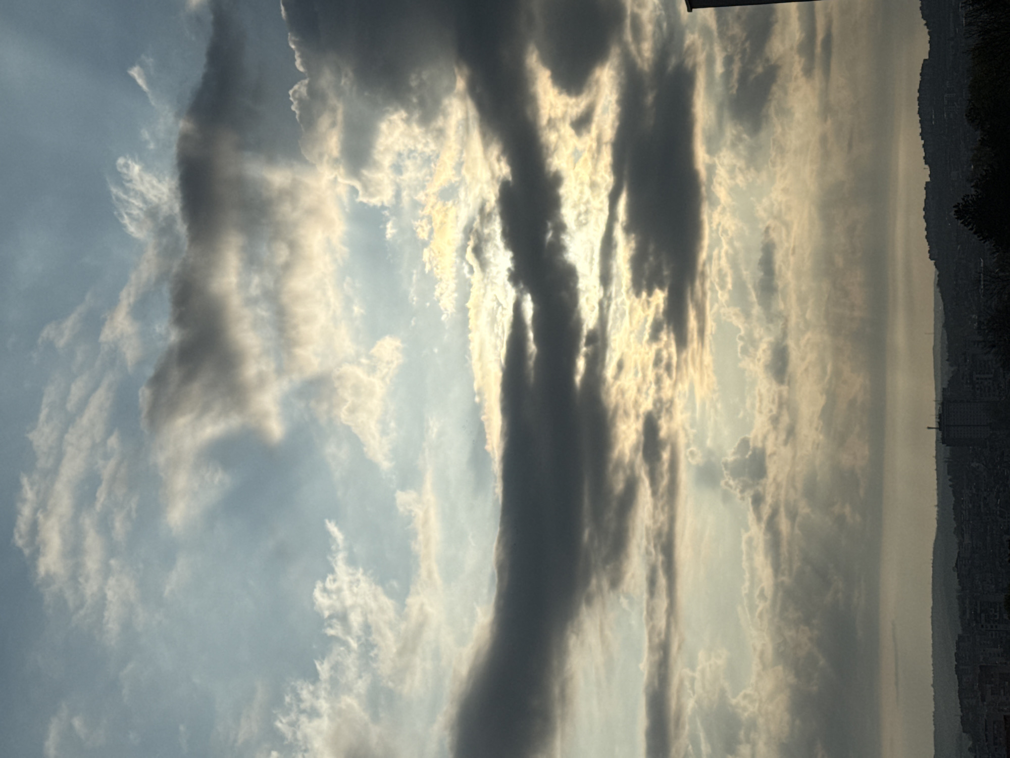 The image is an upward view of the sky, likely taken at dusk or dawn. Wispy, cirrus clouds are scattered across a blue sky, with thicker, darker clouds arranged vertically in the center. The sun is partially obscured by these central clouds, creating sunbeams that shine through. On the right side of the image, a faint outline of a cityscape is visible, with the silhouette of buildings and trees against the bright sky.
The location is plausibly Pleven, Bulgaria, given the presence of cityscape. I am unable to verify that this picture was taken there without more context clues.