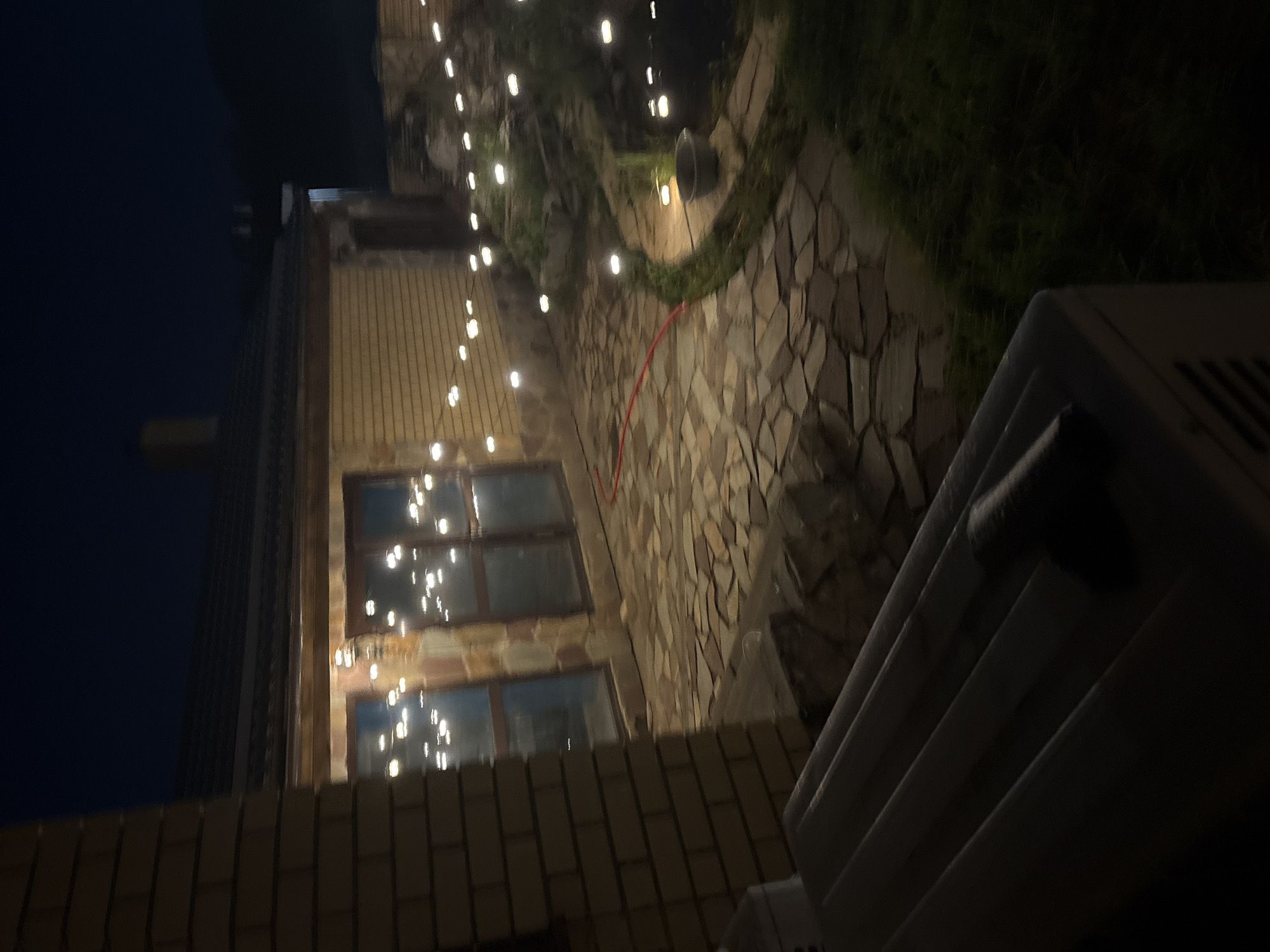 The image depicts a dimly lit outdoor patio area at night, likely in the city of Orekhovka, Russia, given the context. A brick building with large windows, adorned with string lights, is visible. The patio is paved with irregular stone tiles. A red hose is laid across the patio. Green grass and some landscaping elements are also present. In the foreground, a white, ribbed object, possibly an outdoor appliance, is partially visible. The overall scene suggests a residential backyard or outdoor entertainment space during nighttime hours.