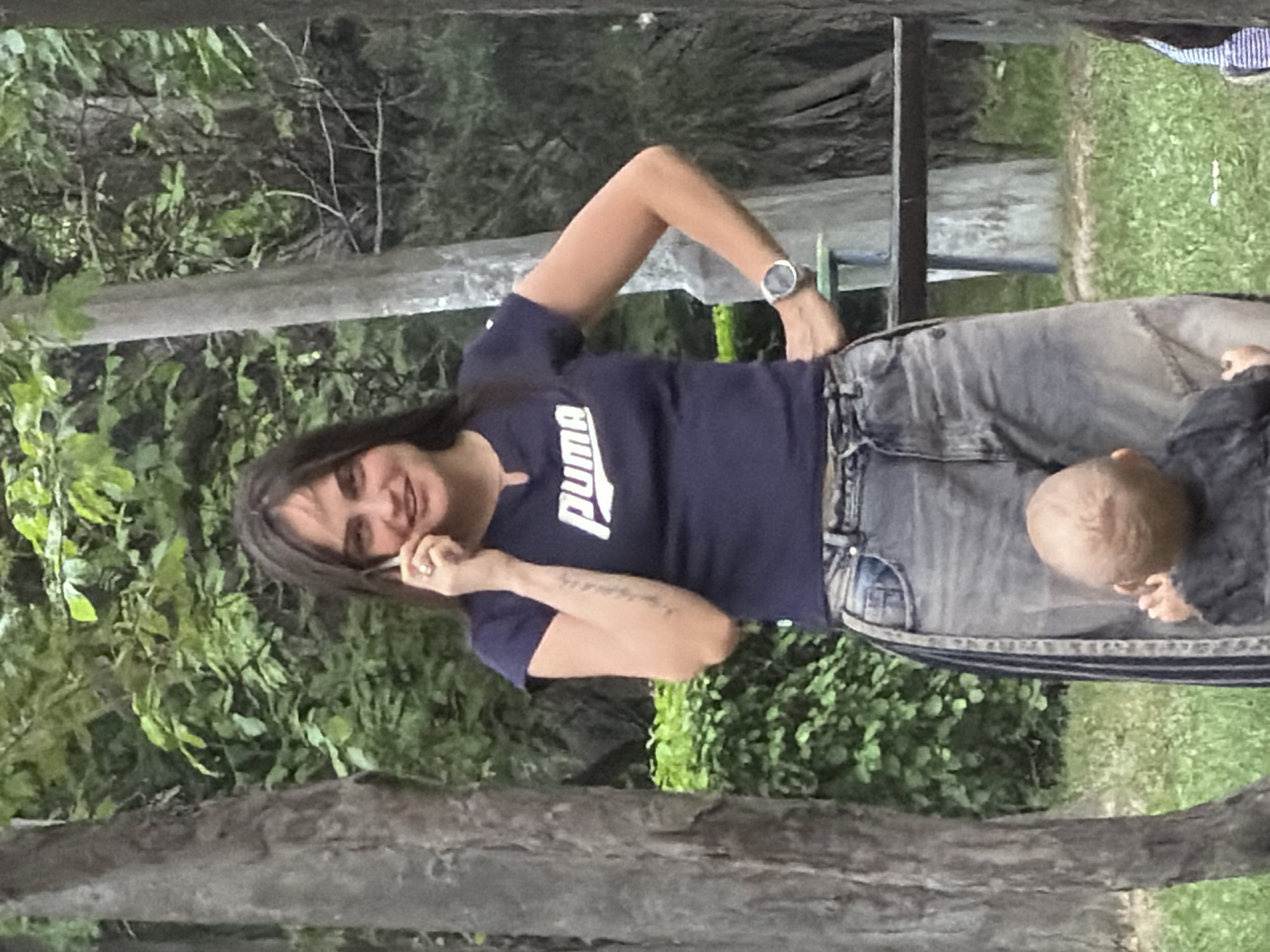In an outdoor, park-like setting with abundant greenery and trees, a woman stands while talking on a mobile phone. She has dark hair and is wearing a dark blue t-shirt with a white Puma logo and loose-fitting denim jeans. A silver watch is visible on her left wrist. In the foreground, a baby wearing a dark fuzzy top is facing away from the camera, with their back to the viewer. The woman's right hand is resting on her hip, and her left arm is bent as she holds the phone to her ear. A faint tattoo is visible on her left forearm. The overall impression is a candid moment captured outdoors.