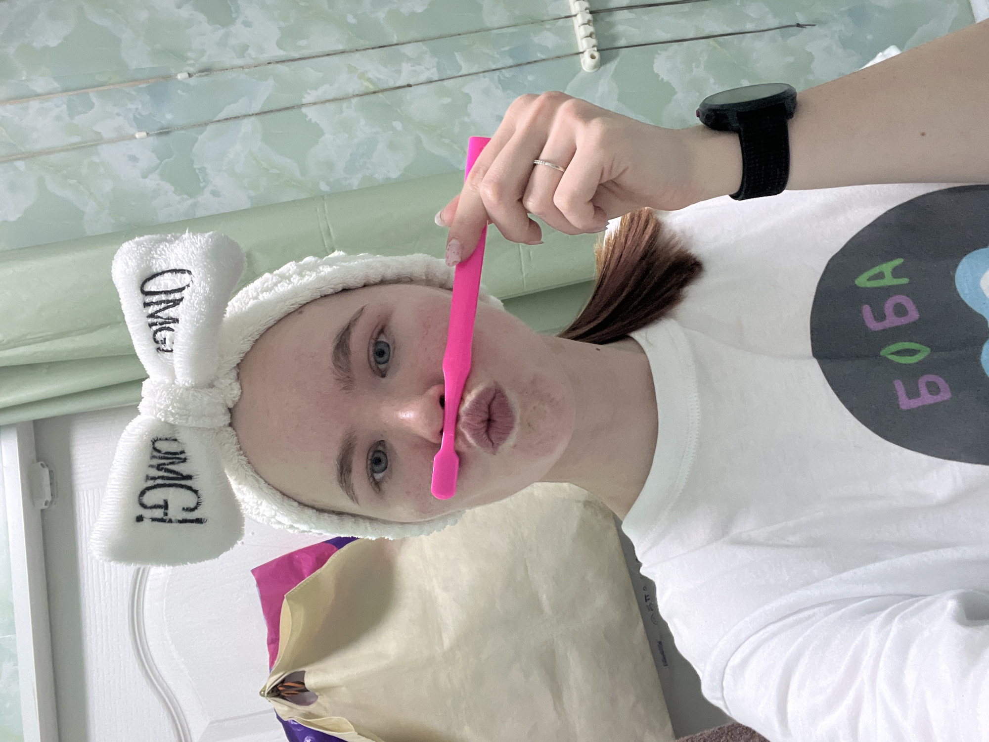A person with fair skin, wearing a white headband with "OMG!" printed on the side and a white T-shirt with "BOBA" in Cyrillic script, is holding a pink tool vertically in front of their lips. The background features a textured wall in light green and white hues, and part of a white door frame is visible on the left. A black smartwatch is on the person's left wrist, and a ring is visible on their left hand. The photo appears to be taken indoors, possibly in a domestic setting, but there is no definitive evidence in the image to confirm its location in Chaikovsky, Russia.