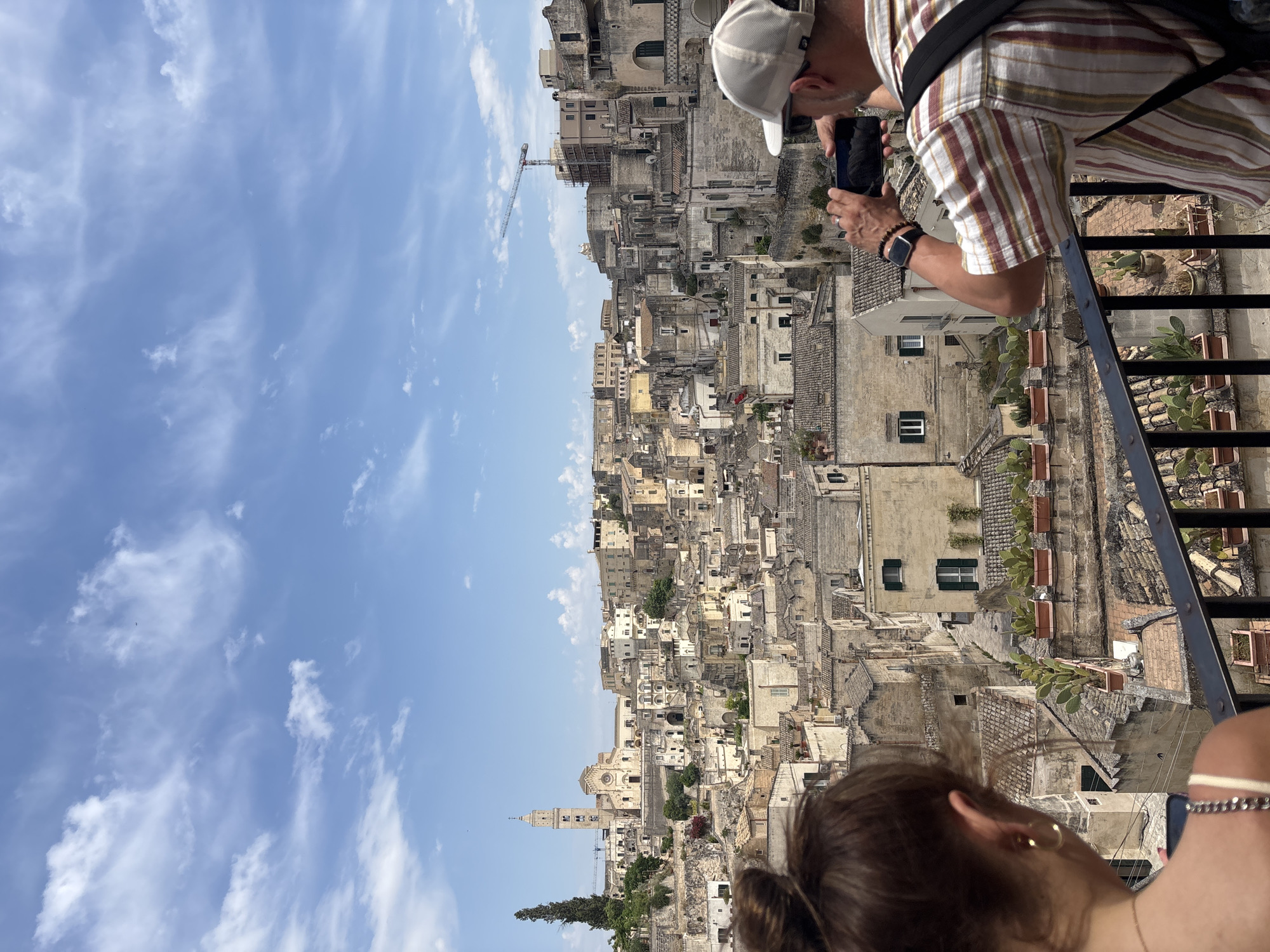 The image, rotated 90 degrees counter-clockwise, presents a panoramic view of a densely populated historic townscape under a bright blue sky with scattered white clouds. In the immediate foreground, a dark metal railing separates the viewer from the scene. To the right, a man, seen from the back and right side, is holding a black smartphone horizontally, appearing to photograph or video the view. He wears a light-colored baseball cap, a striped short-sleeved shirt, and a black backpack. A dark-faced smartwatch is visible on his left wrist. To the bottom left, the back of a woman's head and shoulder are visible; she has brown hair, is wearing earrings and a necklace, and is also looking out at the vista.
The cityscape below consists of numerous old, light-colored stone buildings with a mix of flat and terracotta-tiled roofs, densely packed and seemingly built into a hillside or valley. The architecture suggests an ancient European settlement. Many buildings feature green-shuttered windows. In the distant background on the left, a prominent church or cathedral bell tower rises above the structures. A construction crane is visible in the mid-ground, indicating ongoing work amidst the historic buildings. Rectangular plant pots containing succulents, possibly cacti, are placed along ledges in the foreground buildings. The scene is illuminated by bright daylight, suggesting a clear and sunny day. No discernible text is visible in the image.