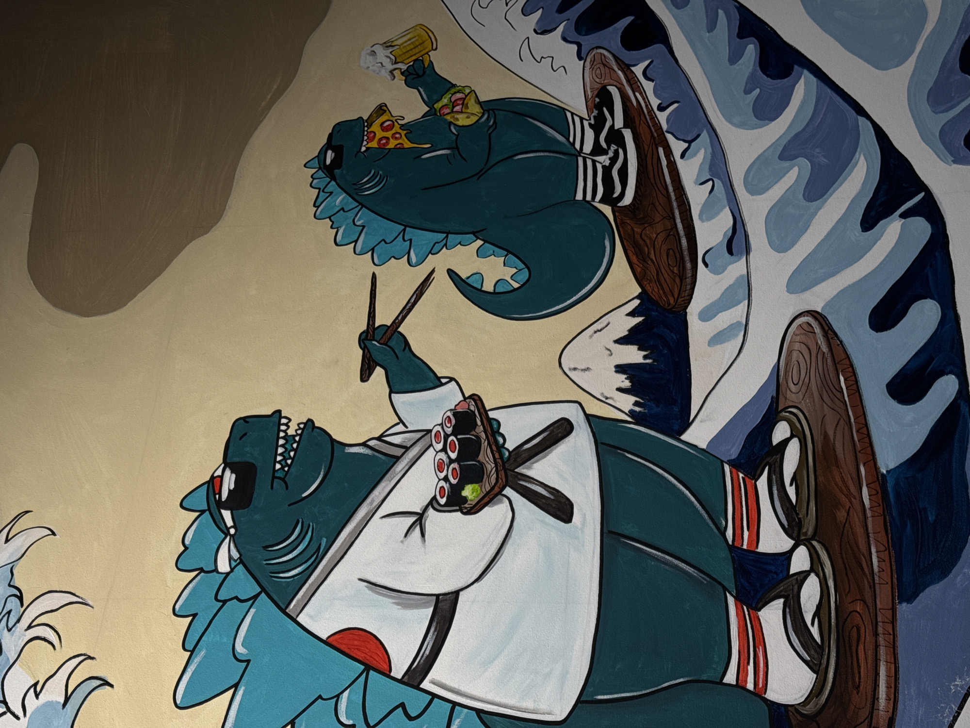 The image is a mural painted on a wall, depicting two cartoon Godzilla figures. The figure on the left is larger and wears a white gi with a red circle on the chest, holding a plate of sushi and chopsticks. It stands on a wooden surfboard amidst stylized blue waves. The figure on the right is smaller, wearing sunglasses, holding a slice of pizza and a taco, and drinking from a beer mug. This figure is also on a wooden surfboard, riding the waves. Behind them, there are stylized mountains and a yellowish-beige background with abstract brown shapes. The mural appears to be located on a exterior wall. There is no discernible information in the image that would indicate its location in the city of Mykolaiv, Ukraine.