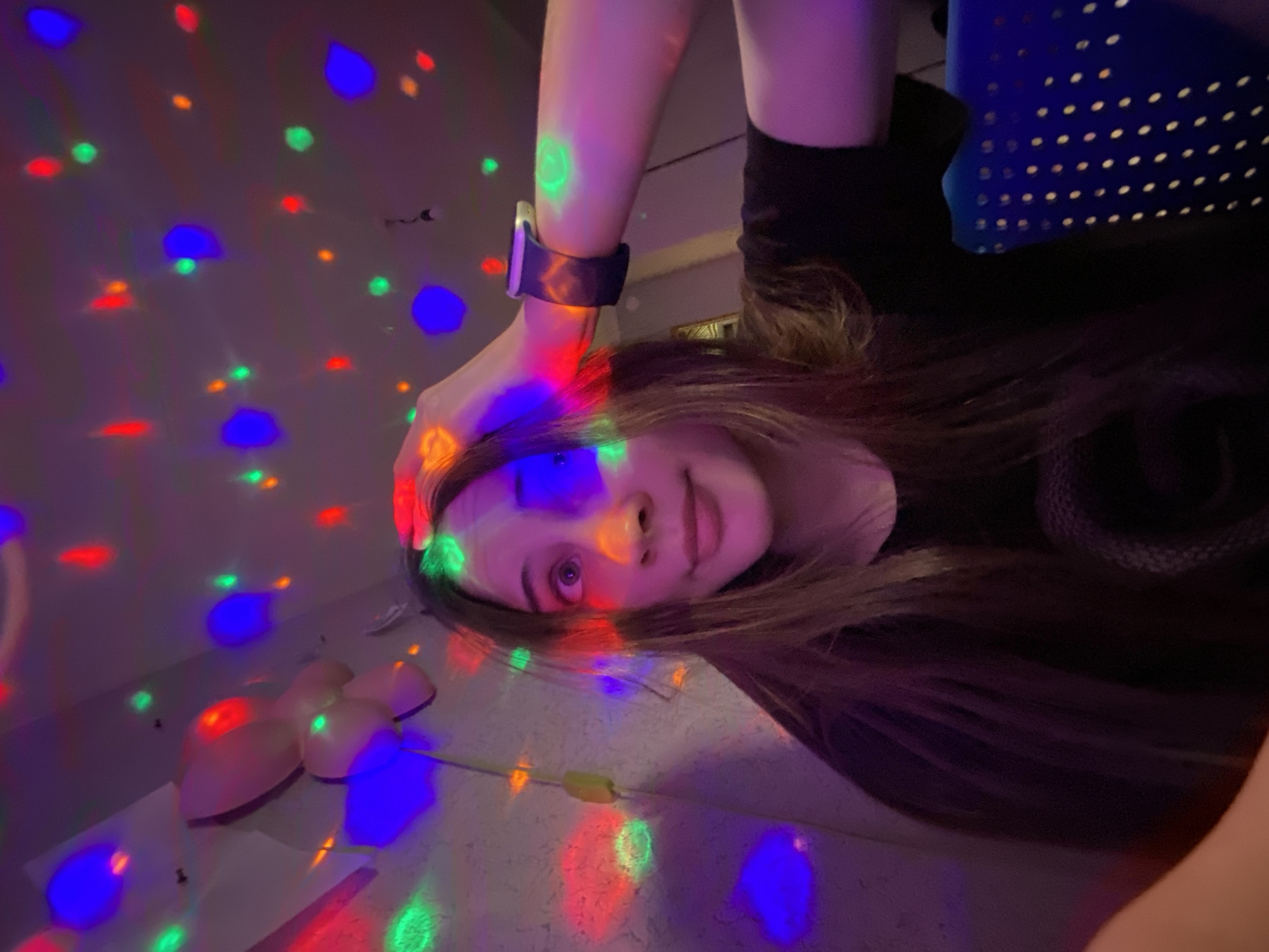 A young woman is visible in the image, illuminated by a multi-colored light source. She is wearing a dark shirt and an electronic watch on her left wrist. A blue, dotted object is near the top right. The room is illuminated with dots of red, blue and green lights. There's a device with a cable on the left. The location is not identifiable as Votkinsk, Russia.
