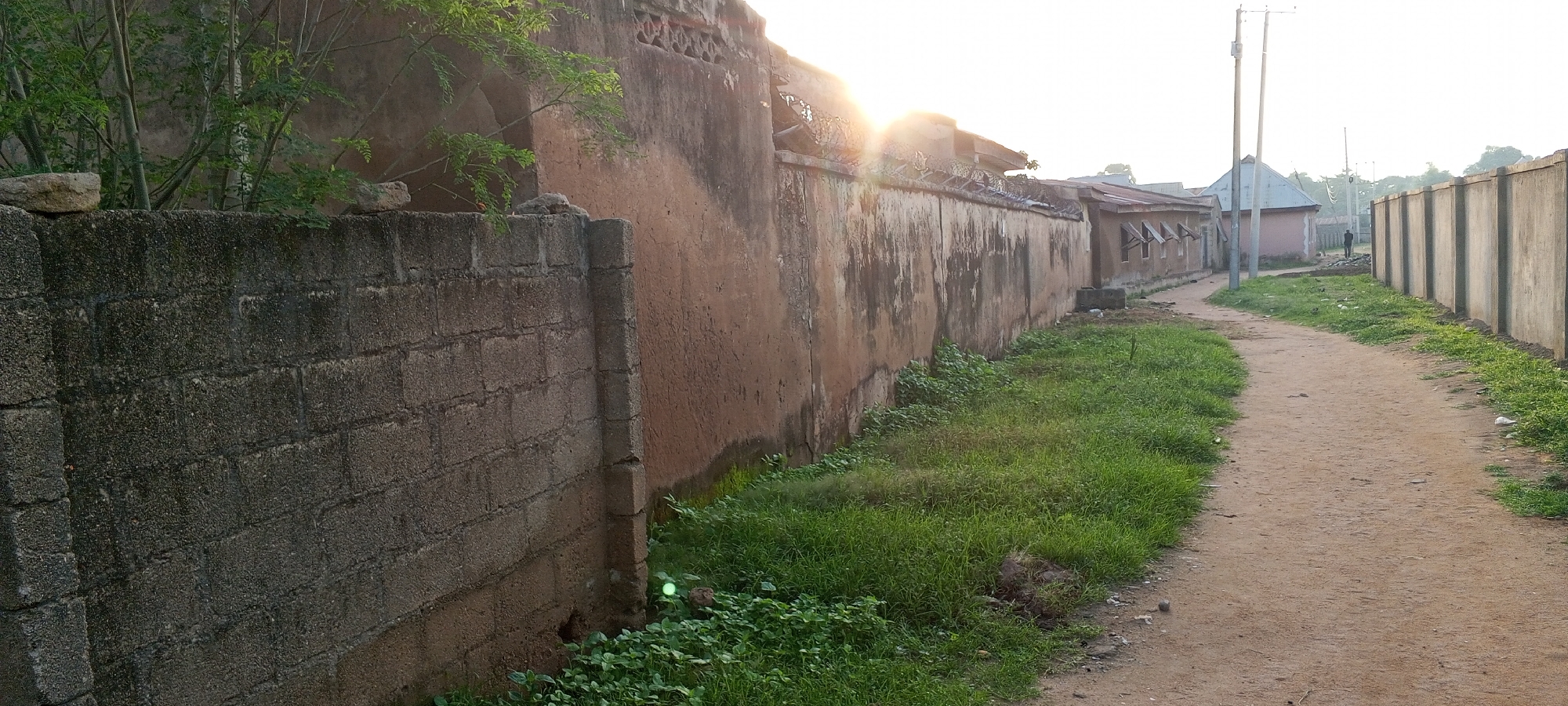 The image depicts a dusty, unpaved road in Jalingo, Nigeria. The road is bordered on the left by a low, grey concrete block wall and on the right by a tall, weathered, light brown plaster wall. Green grass and some small plants grow along the base of both walls. The plaster wall on the right appears to have some decorative elements near the top, including a section with a perforated pattern and barbed wire on top. Behind the plaster wall, a row of buildings with corrugated metal roofs is visible, some with small, awning-covered windows. Further down the road, a person is walking away from the camera. The sky is bright and hazy, suggesting either early morning or late afternoon light. The overall scene conveys a sense of an unpaved, semi-urban or rural environment.
