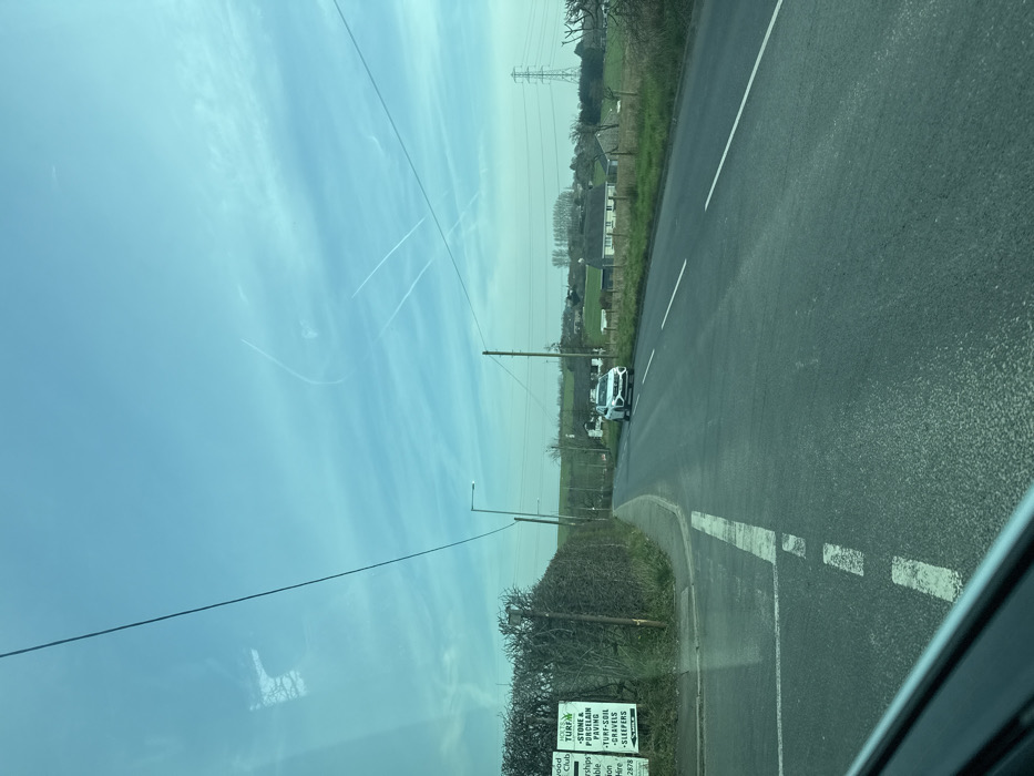 The image displays a two-lane asphalt road under a bright blue sky with scattered cirrus clouds and visible aircraft contrails. The scene is located in a rural or semi-rural area of Bolton, United Kingdom.
In the foreground, the image is framed by the interior of a vehicle, visible in the lower left, with reflections of tree branches on the windshield. The road features white dashed center lines and a solid white line marking the left edge, curving slightly to the left.
On the left side of the road, a dense green hedge runs parallel to the roadway. Behind the hedge, a white sign with green lettering is visible, identifying "Bolton Turf Farm" and listing services including "TURF," "TOP-SOIL," "STONE & PORCELAIN PAVING," "GRAVEL," and "SLEEPERS." A utility pole with overhead wires and a street light pole are positioned along this side.
Ahead on the road, a white SUV, possibly a Hyundai Tucson based on its grille design, is parked or slowly approaching, facing the camera, situated in what appears to be the opposite lane. Its headlights are off.
The background consists of undulating green fields, several scattered trees, and a few detached residential or farm buildings. A prominent large electricity pylon stands on the far right horizon.