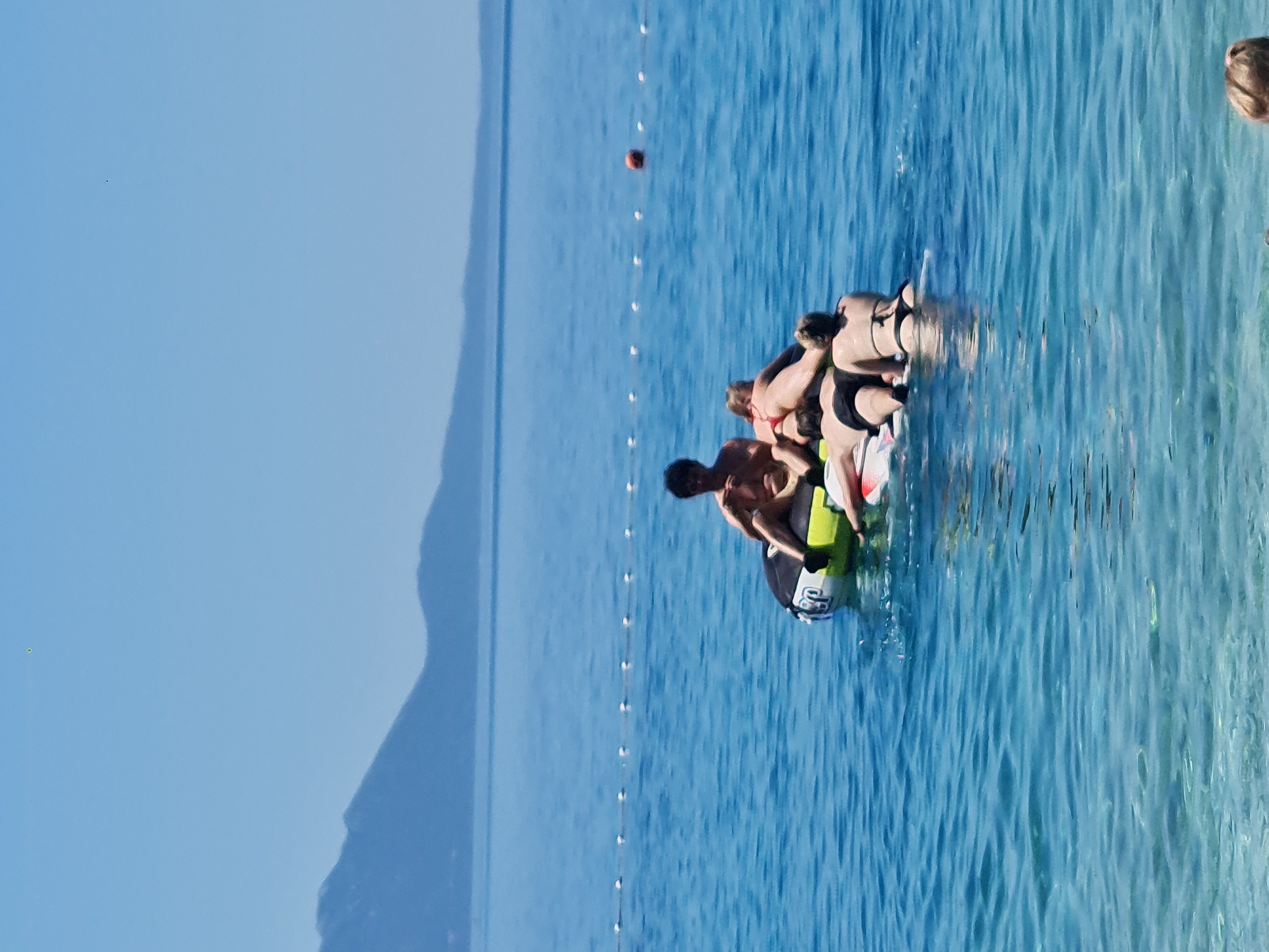 A group of four individuals are on a circular inflatable in the sea, with a mountain range visible in the background under a clear blue sky. One person, a male, is sitting on the inflatable, while the other three, two females and one male, are lying on top of it. The inflatable is partially submerged in the turquoise water. A line of white buoys stretches across the water in the mid-ground, and an orange buoy is visible further out. The scene is set in Općina Gradac, Croatia, a coastal town on the Makarska Riviera.