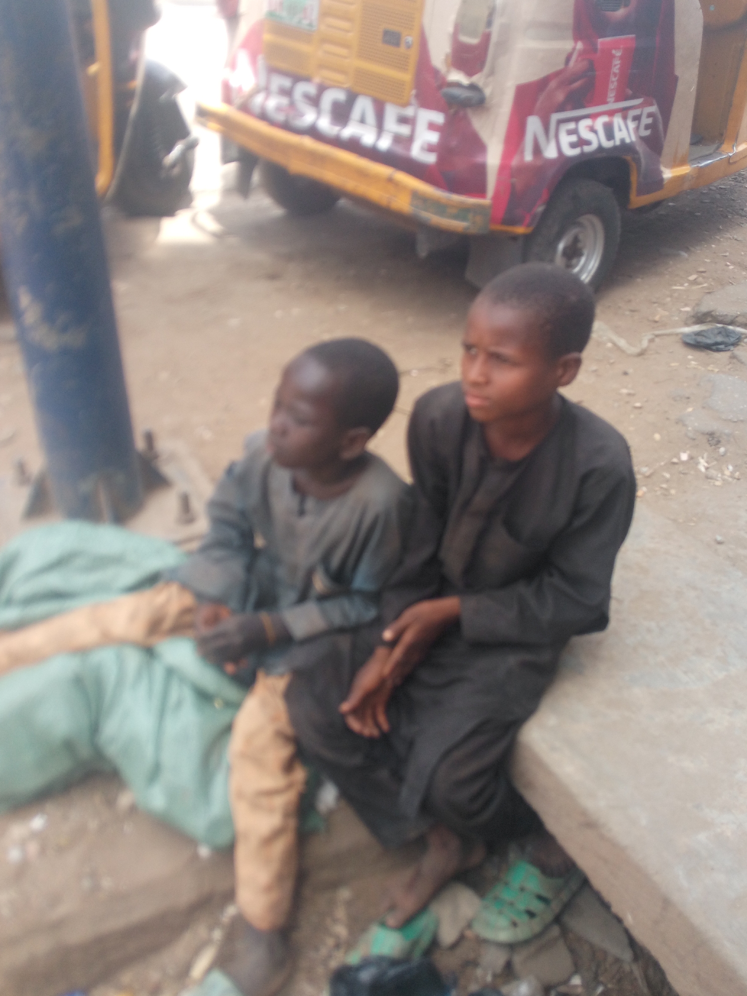 Two young boys are sitting close together on a concrete structure. One boy wears a gray tunic and tan pants, while the other wears a dark robe or tunic and green sandals. They both have dark skin and short hair.
Behind them is a motorized tricycle, which is yellow and decorated with a large "NESCAFE" advertisement. The environment appears to be an urban street in Maiburiji, Nigeria, with a pole located on the left side of the image. Debris and litter are visible on the ground.