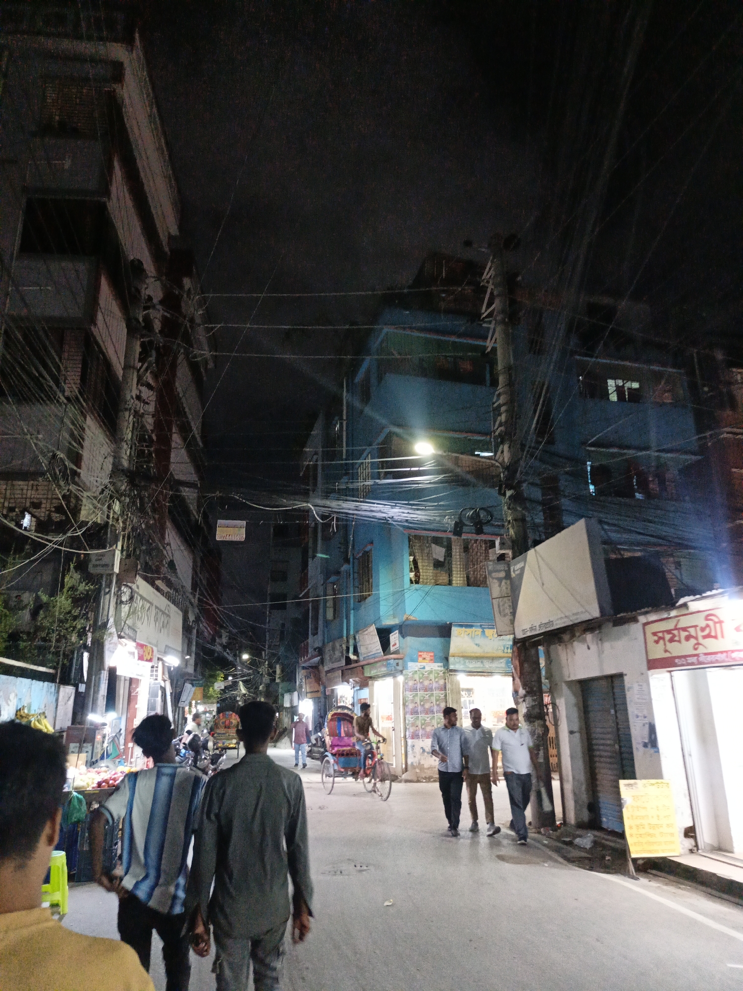 The image depicts a narrow, bustling street scene at night in Dhaka, Bangladesh, likely in a densely populated commercial or residential area characterized by multi-story buildings. The foreground shows the backs of three young men walking away from the camera down the street. The man on the left wears a blue and white striped shirt, the middle man a dark gray shirt, and the man on the right a light gray shirt. They are on a paved street, which is illuminated by overhead lights and lights emanating from shops and buildings.
The middle ground of the image reveals more activity. A cycle rickshaw, a common mode of transport in Dhaka, is visible with passengers and a driver. To the left of the street, there is a fruit stall with various fruits displayed. Further down the street, more people are seen walking and interacting. The buildings lining the street are close together, with many windows and balconies visible. A prominent feature of the urban landscape in Dhaka, the dense network of electrical wires and cables, crisscrosses overhead, obscuring the night sky in many areas. Some buildings have signs with Bengali script. The overall impression is one of a vibrant, active urban environment at night.