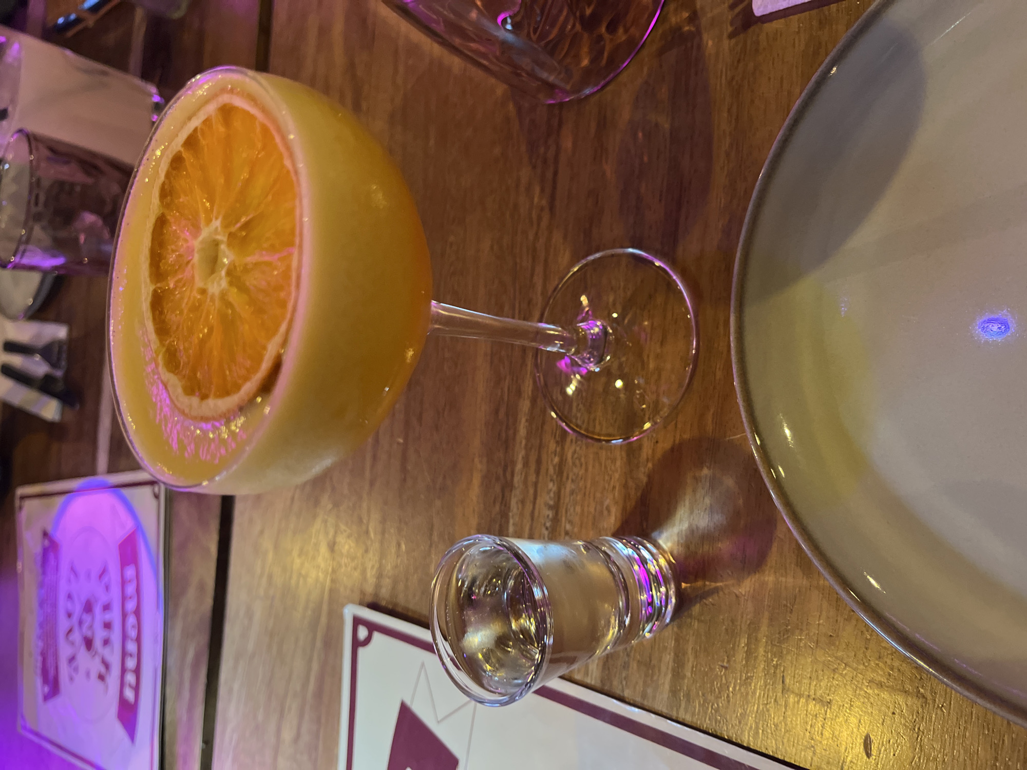 The image depicts a cocktail, possibly a mimosa, served in a coupe glass, garnished with a thick slice of an orange. A shot glass filled with a clear liquid sits near the base of the coupe glass. A plate is visible to the right, partially in the frame. Menu cards with purple and white details are also visible on the wooden table. The location of the image is Saint-Jean-de-Védas, France.