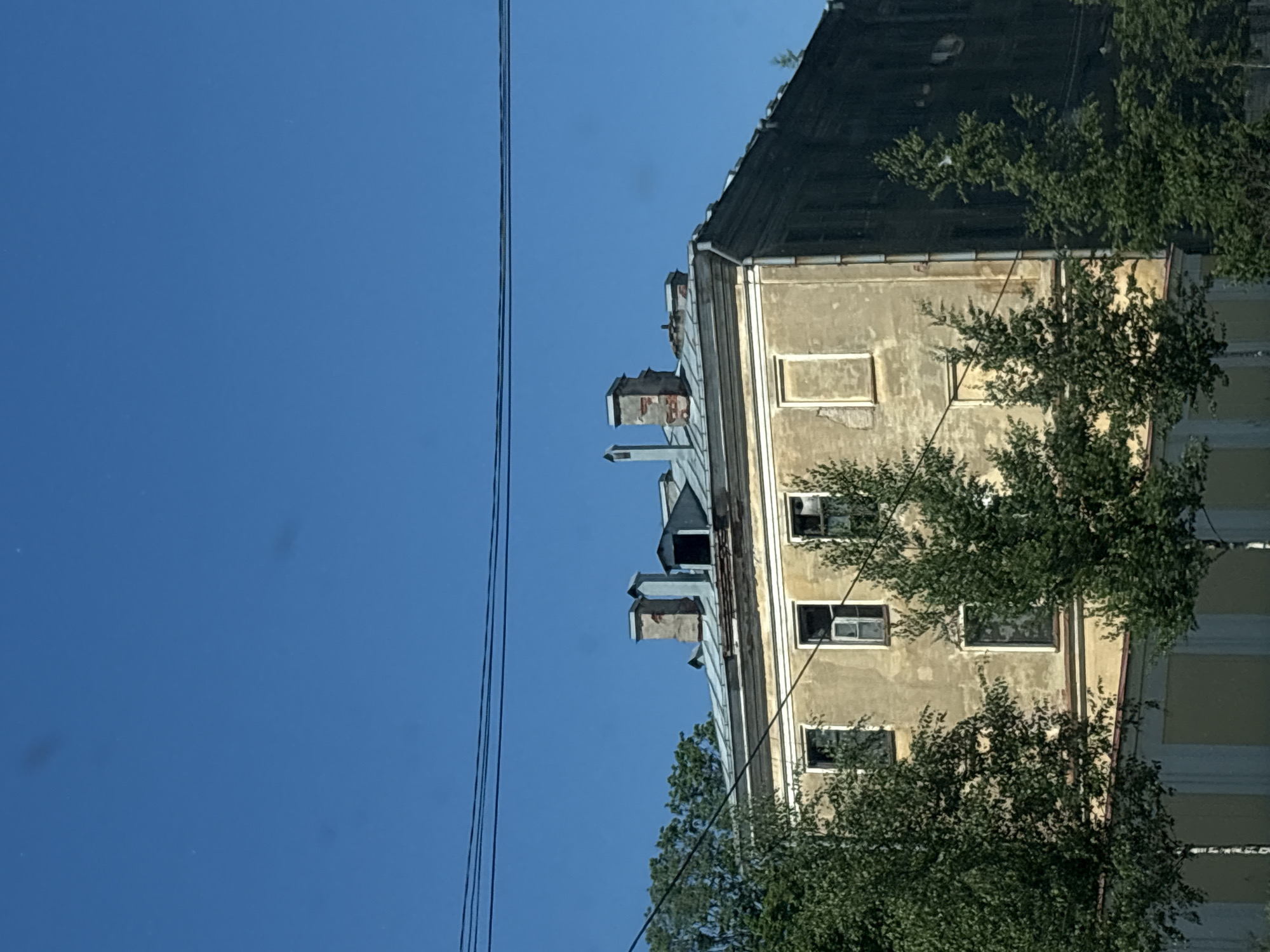 The image shows an old, weathered building in Saint Petersburg, Russia. The building is multi-story with a light yellow facade and a dark, aged roof. Several windows are visible, some with broken panes, and some are covered or boarded up. The roof features multiple chimneys and skylights, indicating an older architectural style. Trees with lush green foliage partially obscure the lower floors of the building. Power lines are strung across the upper portion of the image against a clear, bright blue sky. The overall impression is one of an aged, possibly disused building in an urban setting, common in historical districts of Saint Petersburg.