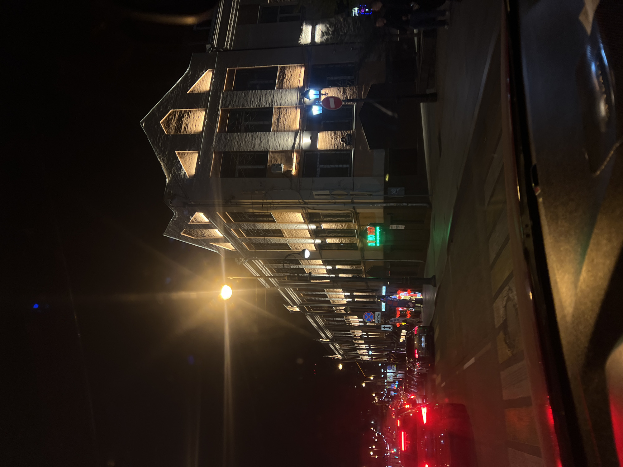 The image shows a street scene in Vladivostok, Russia at night. The view is from inside a car, looking out at a building facade illuminated with warm light, with a pedestrian crossing in front of the building. Further down the street, there is traffic with visible red tail lights, and street lights. There are street signs visible, along with neon signs and other urban infrastructure. People are visible in front of the building.