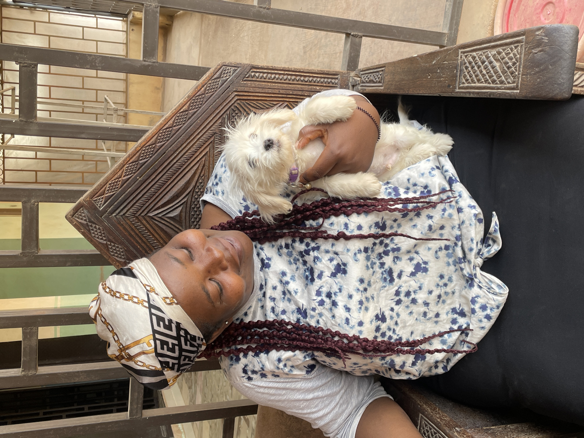 A woman is lying back in a carved wooden chair holding a small white dog, outdoors. The woman is wearing a white and blue floral patterned shirt, light gray shorts, and a white headscarf with a black and gold chain pattern. She has dark hair styled in maroon braids or dreadlocks. The dog, held in her arms, has a purple collar and appears to be looking at the camera. The chair has elaborate carvings on its arms and back. A metal railing is visible in the foreground. The location is Barakin Akawo, Nigeria.
