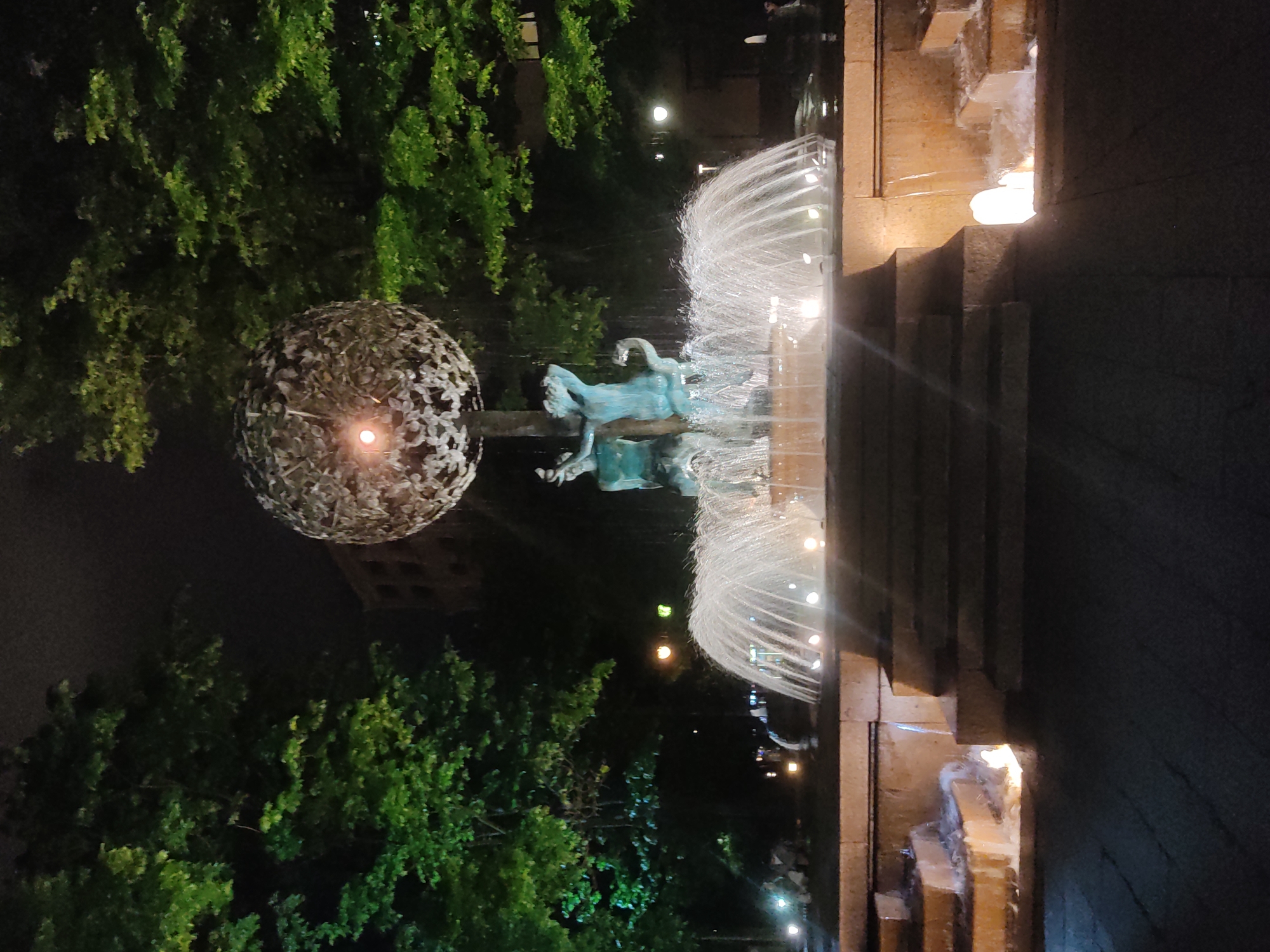 The image shows a nighttime view of a fountain in Moscow, Russia, with a sculpture of figures in the center. The fountain has several tiers of water cascading down, illuminated by lights. To the left, there is a decorative spherical object with a light in the center, surrounded by green trees. The background includes building facades and streetlights.