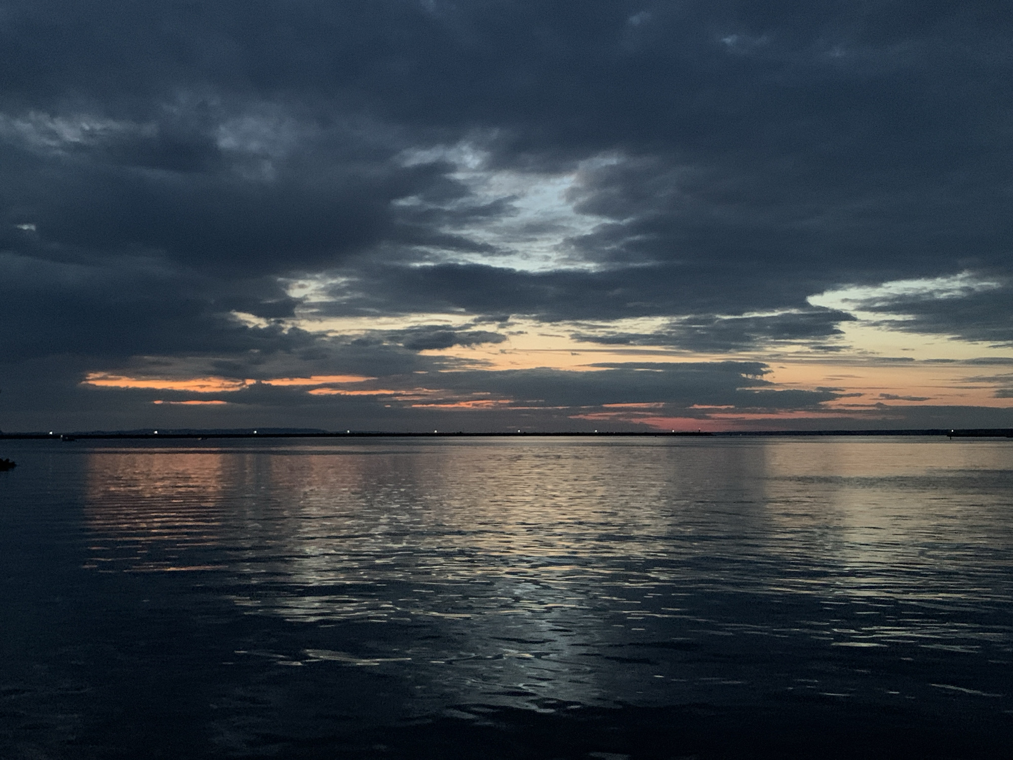 The image depicts a serene twilight scene over a body of water, possibly the Kama River, near Chaikovsky, Russia. The sky is filled with dramatic, dark clouds, interspersed with patches of light blue and hints of orange and pink near the horizon, suggesting the last vestiges of sunset. The water is calm, reflecting the colors of the sky and creating a shimmering effect. A distant, dark shoreline is visible with a few small lights, possibly indicating a town or industrial area. The overall atmosphere is peaceful and slightly melancholic. There are no visible people or specific landmarks that would pinpoint a precise location within Chaikovsky.