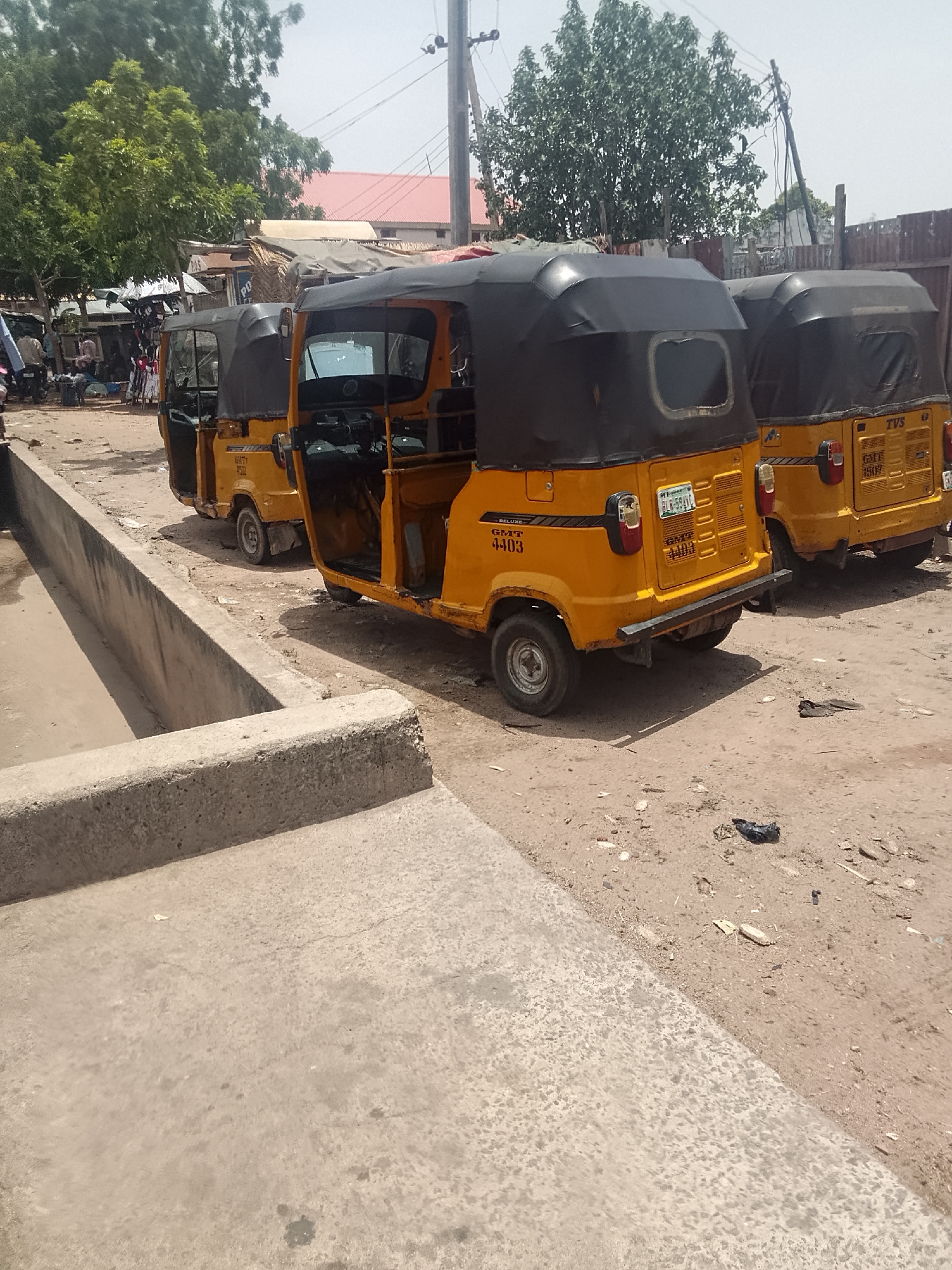 The image shows three yellow auto-rickshaws, also known as "keke," parked on a dusty lot next to a concrete structure, possibly a drainage ditch or raised walkway. The location is likely in Nigeria, given the vehicles' license plate. The auto-rickshaws are the main subjects, parked side-by-side. The middle auto-rickshaw has its driver's side door open. In the background, there are trees and indistinct structures. People are visible in the far left background. The scene is a typical urban transportation setting.